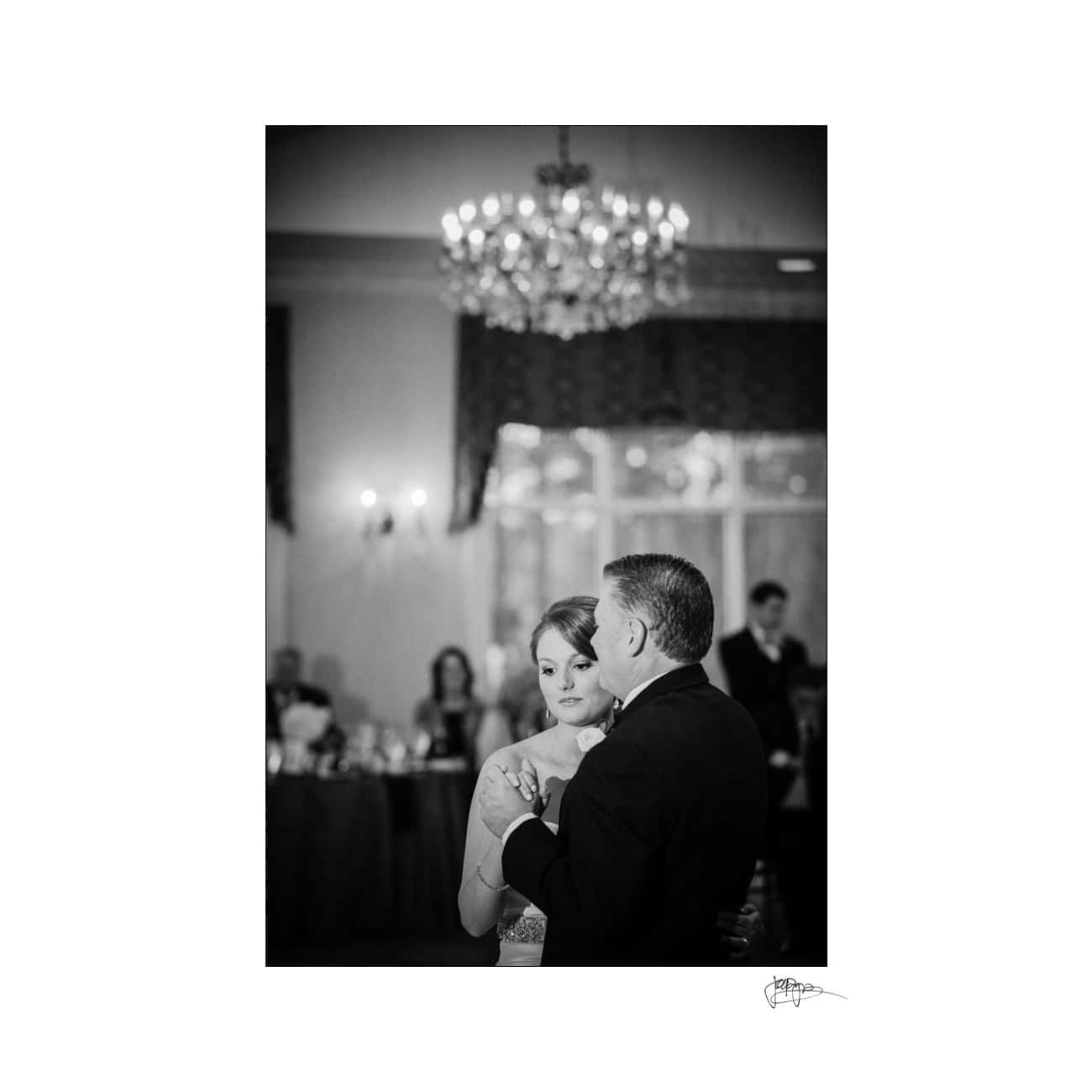 A black-and-white photo of a couple dancing at a wedding reception. The bride wears a strapless dress, and the groom is in a suit. They are holding each other closely, with guests and a chandelier visible in the softly lit background.
