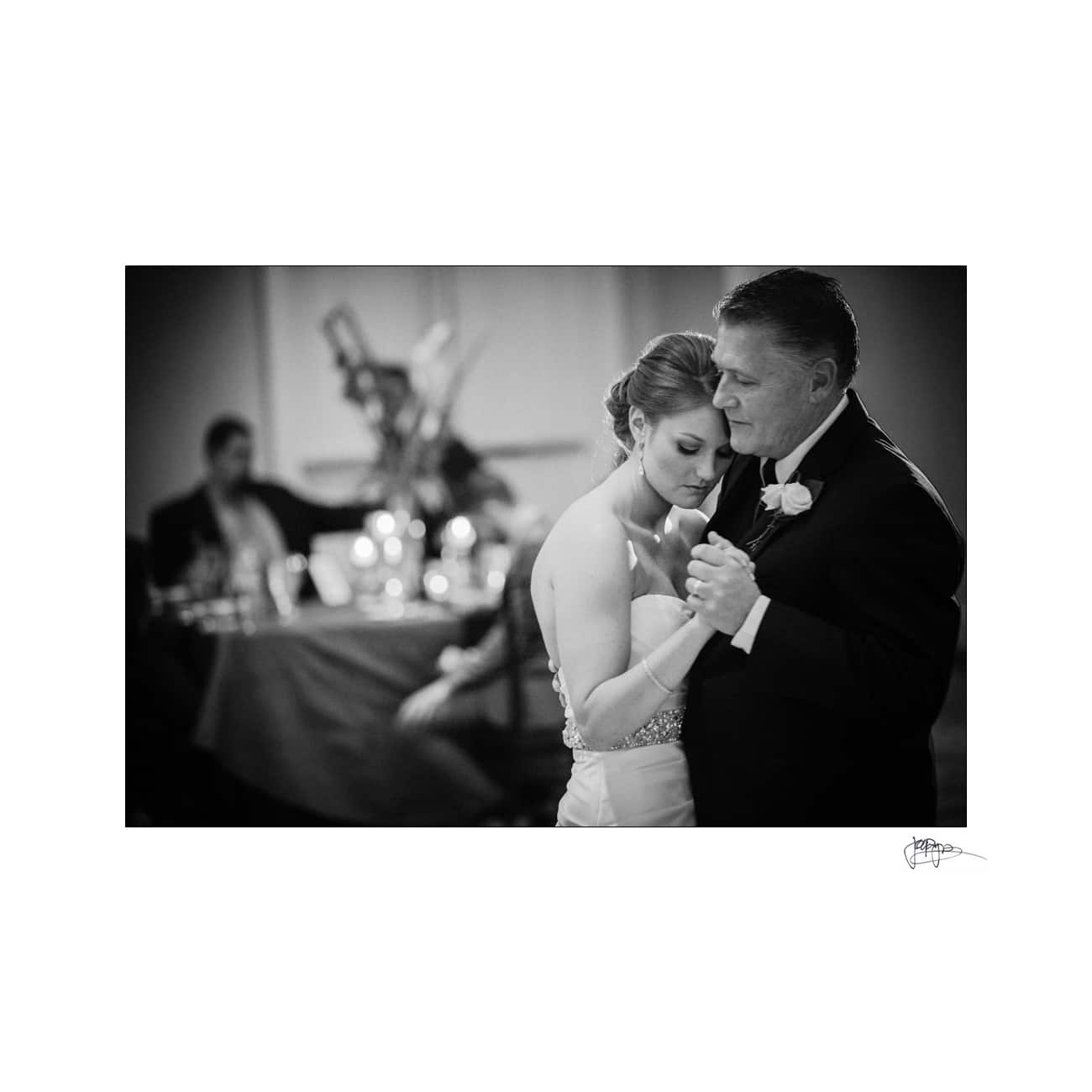 A black and white photo of a bride and an older man sharing a slow dance at a wedding reception. They appear emotional and close. In the background, guests sit at a table with glowing candles.