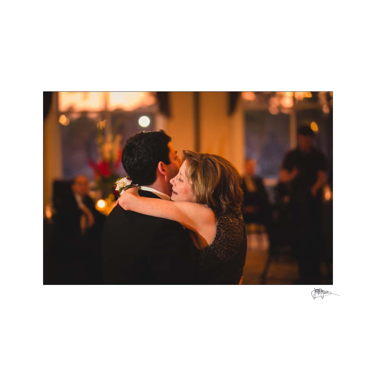 A woman and a man share a close embrace while dancing at a dimly lit event. The woman has her eyes closed, and the background is softly blurred, with people and warm lighting creating a cozy atmosphere.