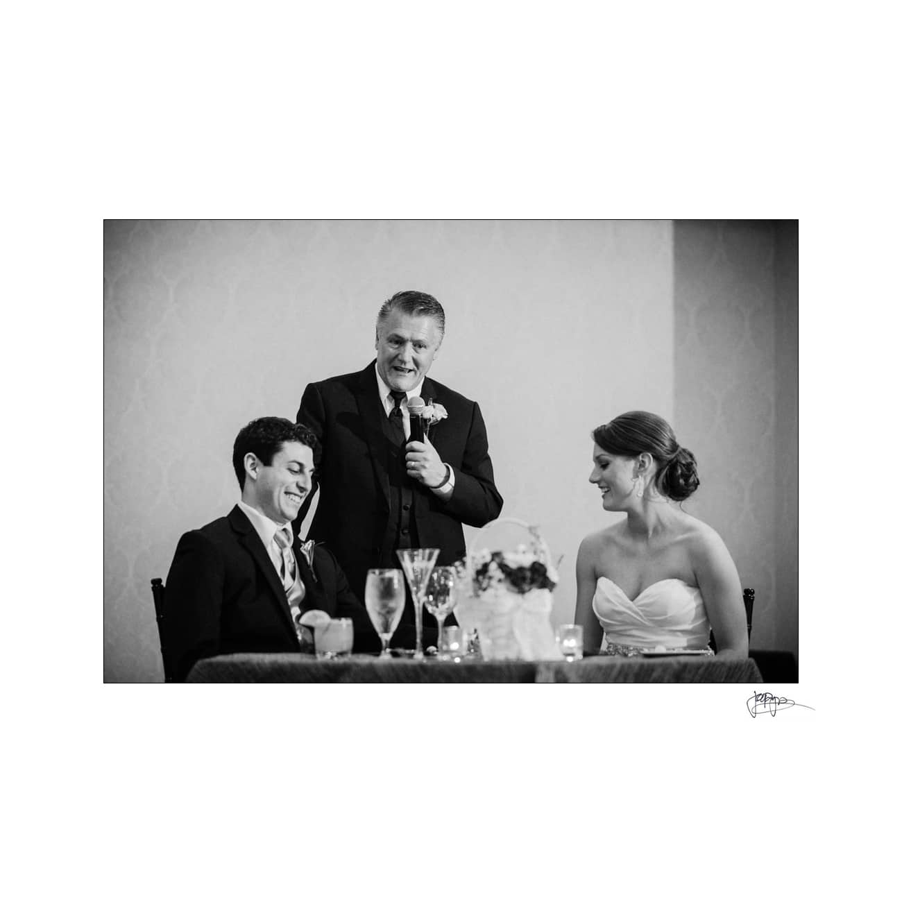 A black and white photo of a wedding reception. A man in a suit is standing with a microphone, smiling and speaking to a seated couple, who are also smiling. The bride is in a strapless gown, and the groom is in a suit. Glasses are on the table.