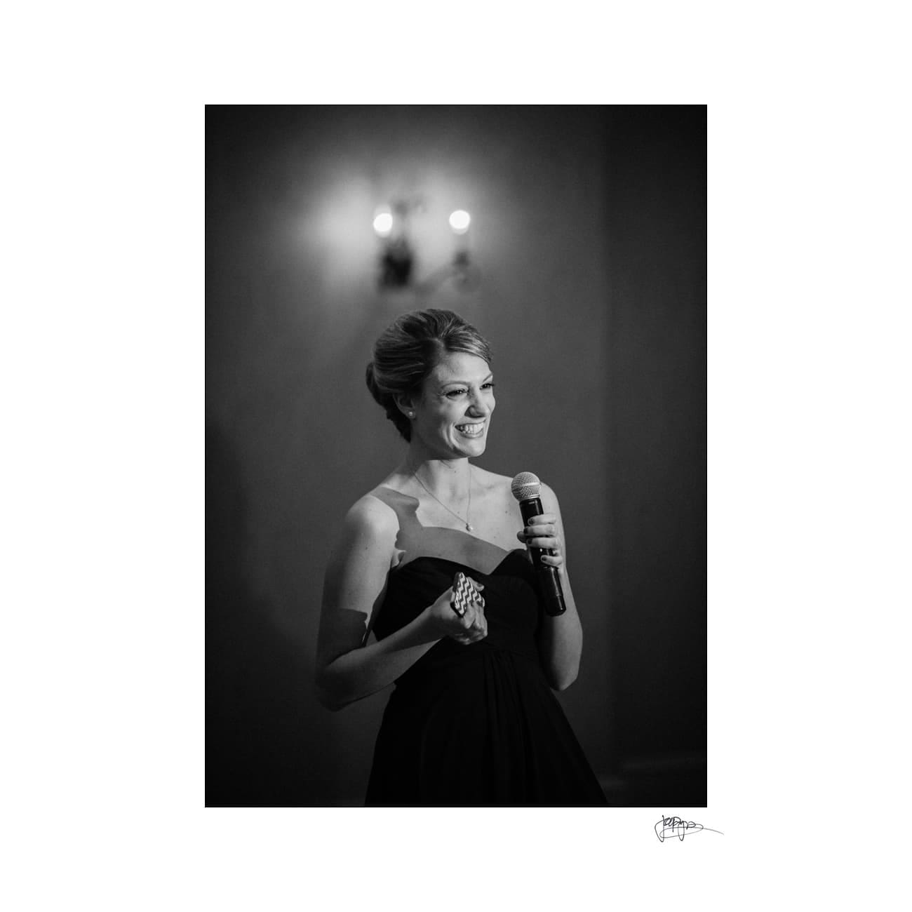 A woman in an elegant dark dress speaks into a microphone, smiling warmly. She stands in a softly lit room, with two blurred wall lights in the background. The image is black and white.