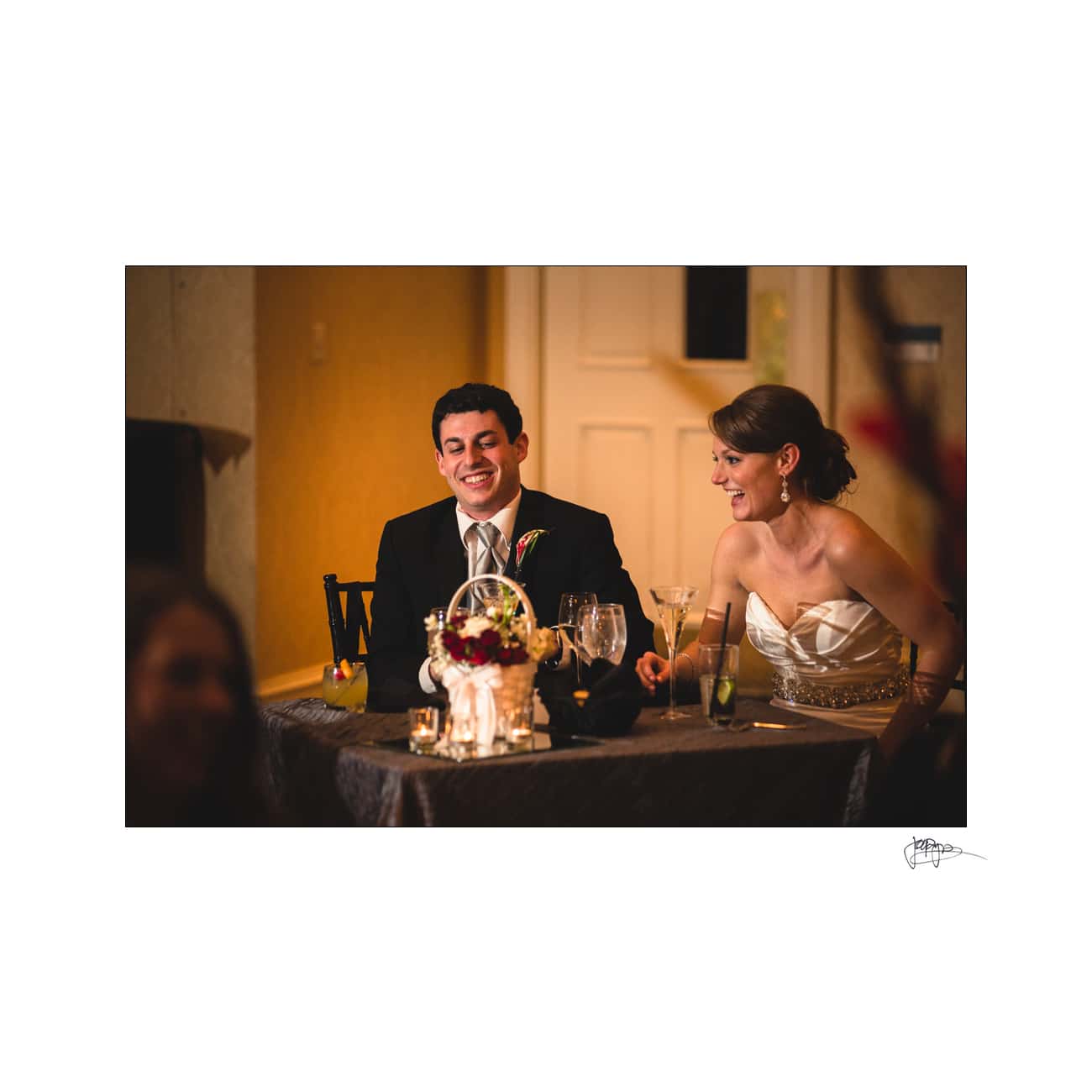 A couple sits at a decorated table during a formal event. The man is wearing a dark suit, and the woman is in a white strapless dress. They are both smiling, surrounded by glasses and a small floral centerpiece.