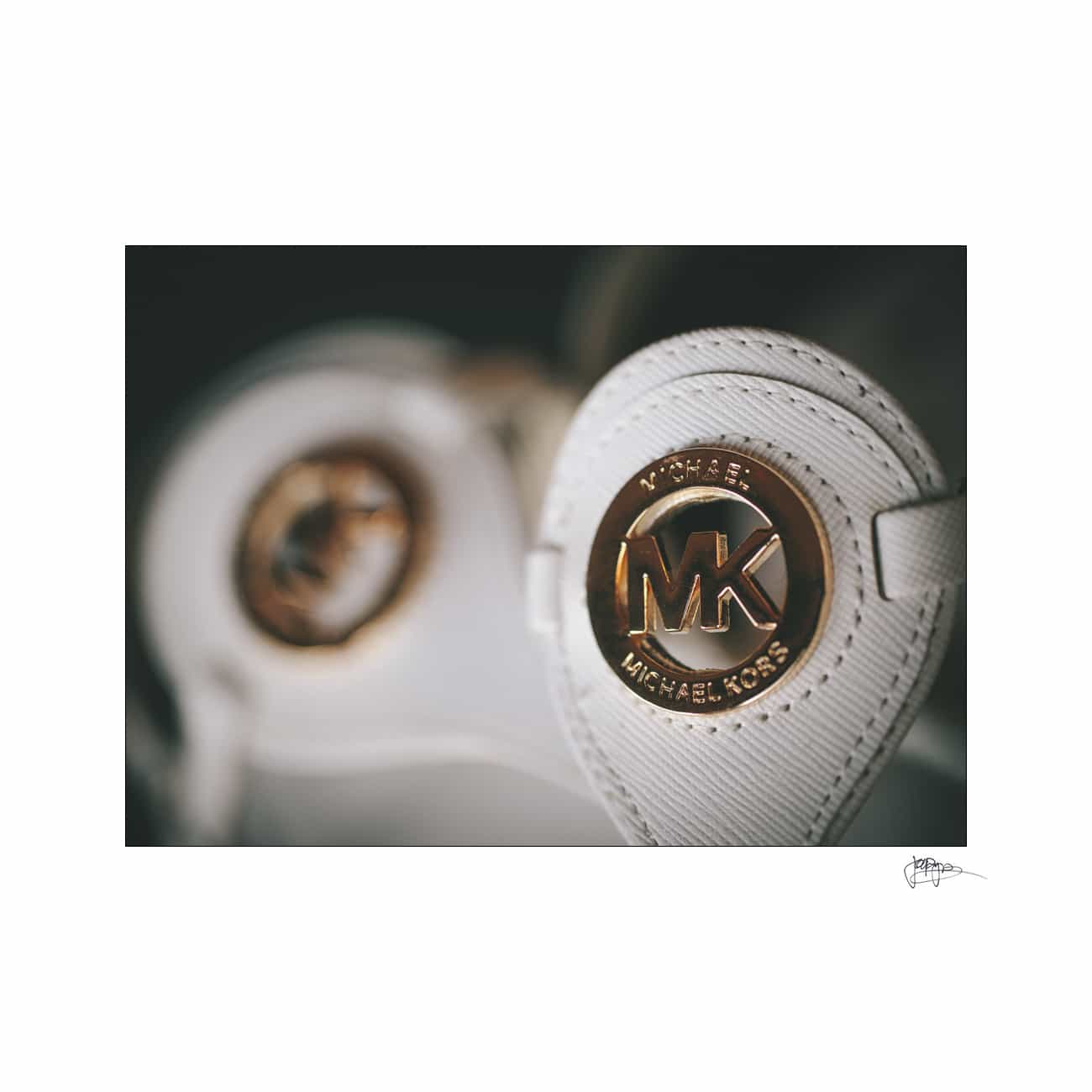 Close-up of a white handbag featuring a gold-colored Michael Kors logo medallion. The logo is embossed with MK and the brand name Michael Kors in stylish font. The background is blurred, highlighting the logo in focus.