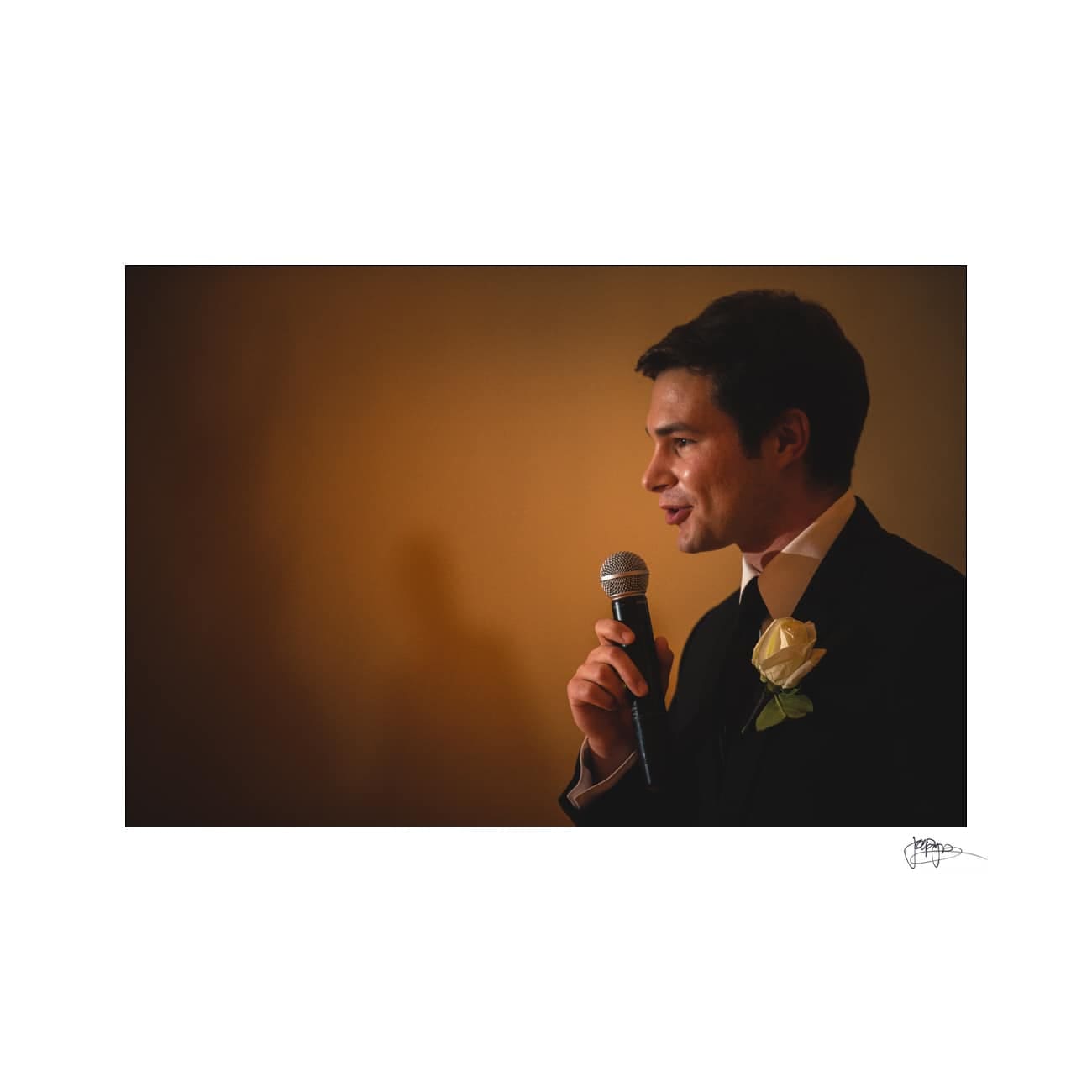 A man in a suit holds a microphone while giving a speech. He has a white rose pinned to his lapel and is standing against a warm, softly lit background.