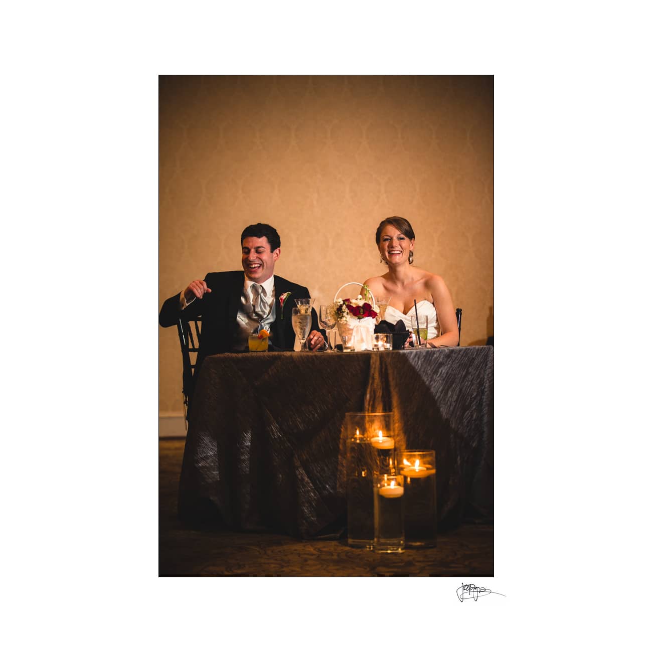 A bride and groom sit at a table during their wedding reception. They are smiling and holding drinks, with flowers and candles on the table. The setting is elegantly decorated with warm lighting.