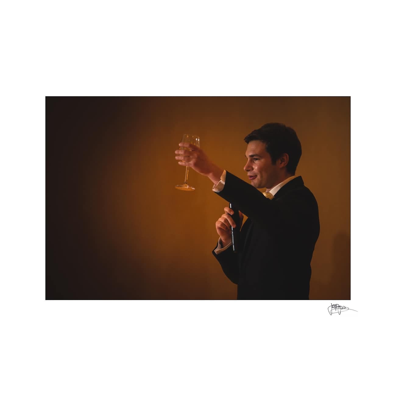 A man in a suit holds a microphone in one hand and raises a wine glass in a toast with the other. The background is softly lit in a warm tone.