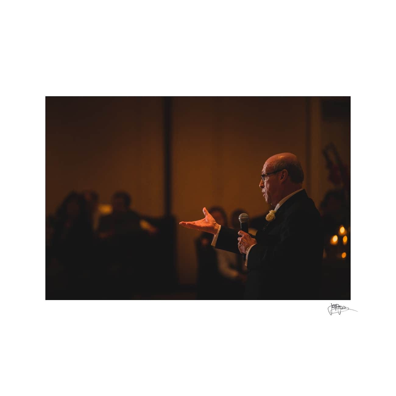 An elderly man in a suit and glasses holds a microphone, delivering a speech with an expressive hand gesture. The dimly lit background hints at an event or gathering, with silhouettes of seated attendees.