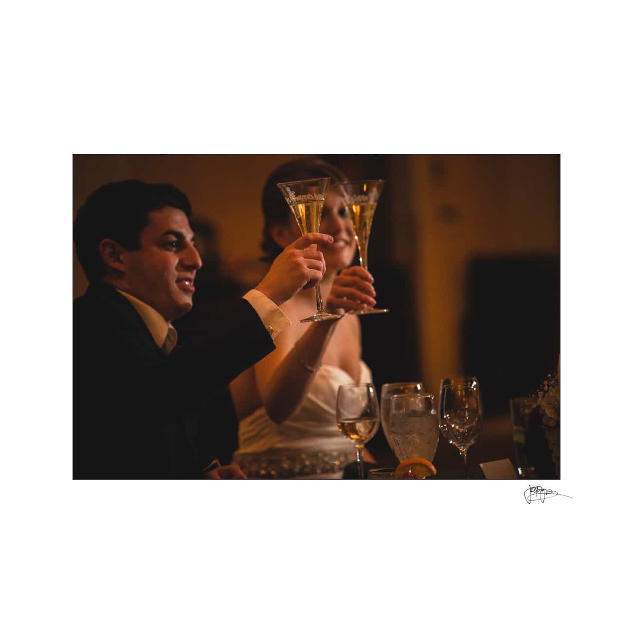 A couple elegantly dressed clinks champagne glasses, surrounded by dim lighting. They appear to be celebrating at a special event. Various glasses and a lemon garnish are visible on the table.
