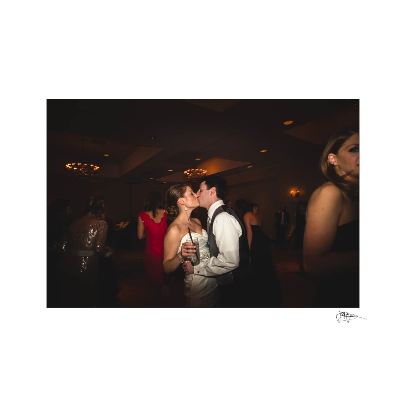 A couple shares a kiss on a dance floor at a formal event. The woman wears a white dress and holds a drink, while the man is in a white shirt and tie. Other guests in evening attire are visible in the dimly lit background.