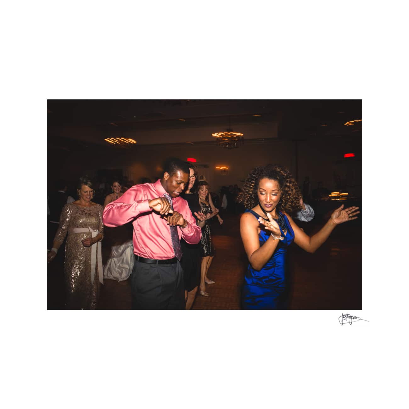 Two people dance energetically in a dimly lit room with other people in the background. The man wears a pink shirt and the woman wears a blue dress. They appear to be enjoying the music and atmosphere.