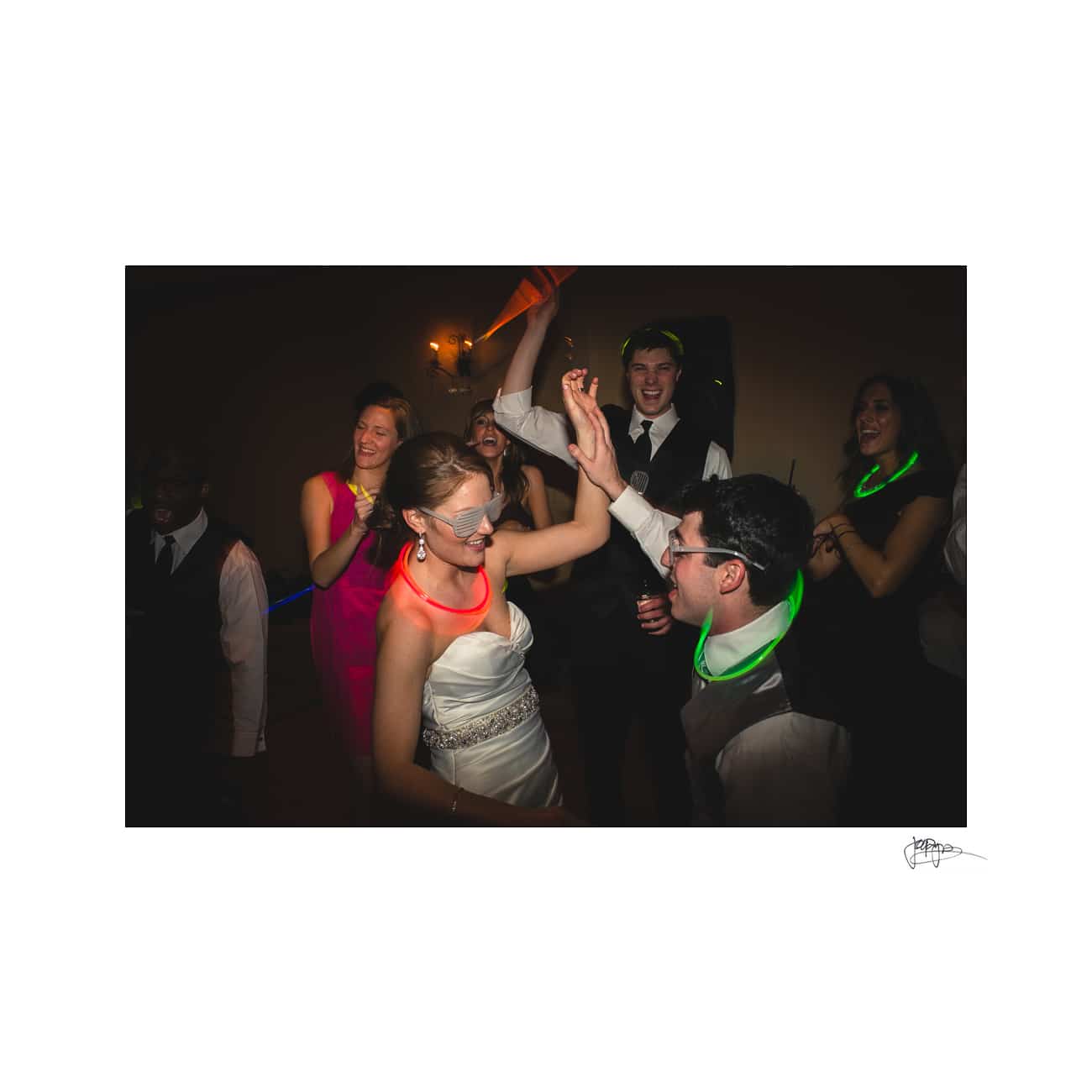 A bride and groom, both wearing glasses and glowing necklaces, dance joyfully with friends at a lively party. Guests surround them, smiling and clapping, with dim lighting and a festive atmosphere.
