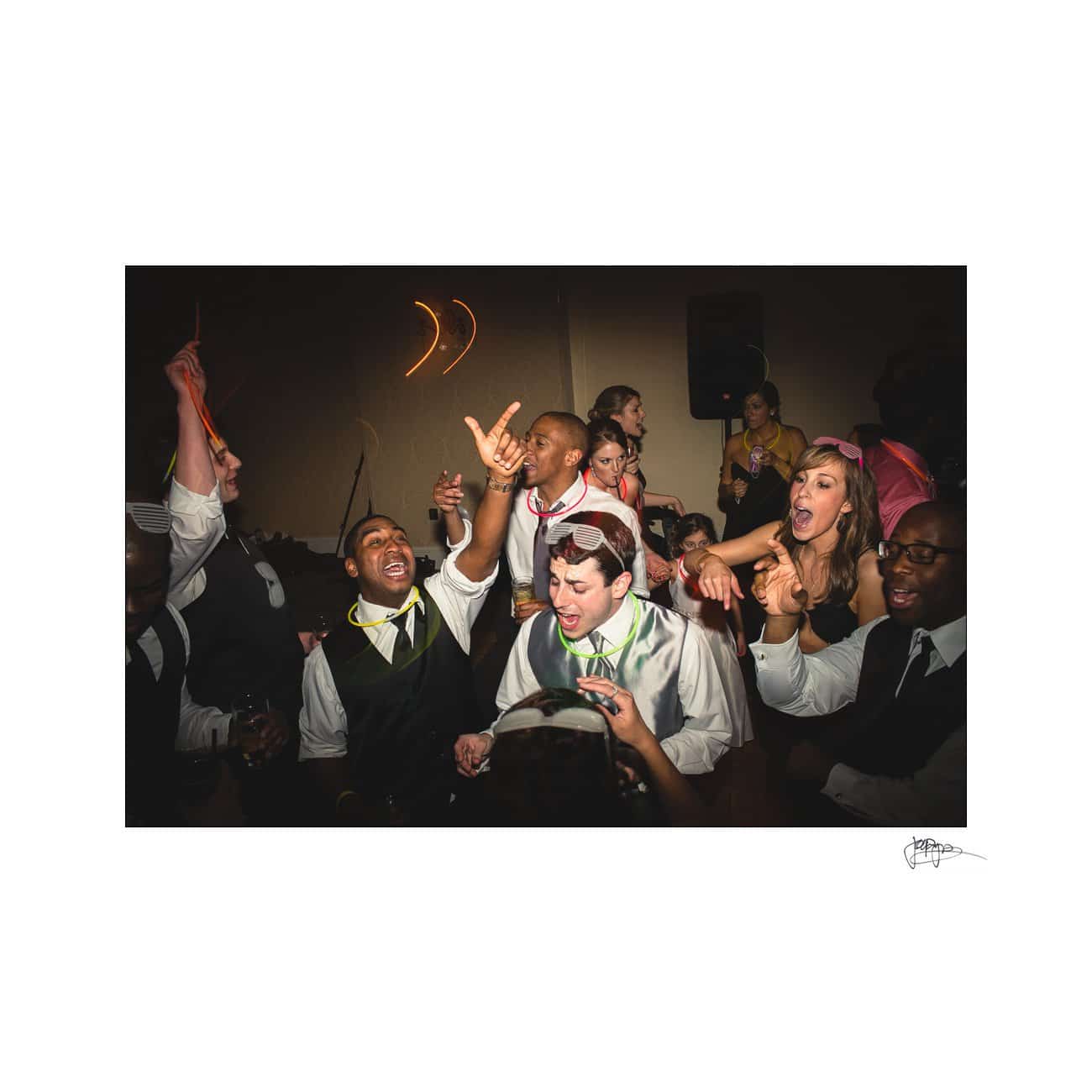 A group of people dressed in formal and semi-formal attire are enthusiastically dancing and cheering at an indoor event. Some hold drinks and wear accessories like sunglasses. The atmosphere is lively and joyful.