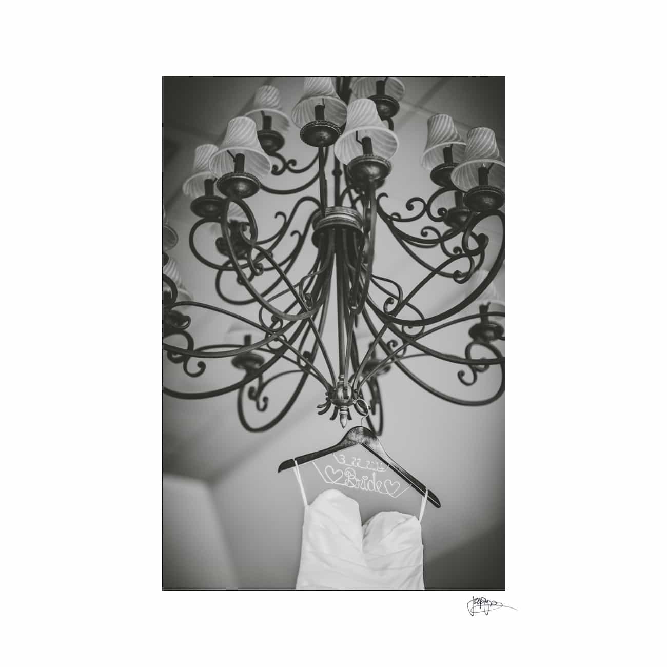 Black and white photo of a strapless wedding dress on a hanger with Bride written on it, hanging under an ornate chandelier with multiple light fixtures.