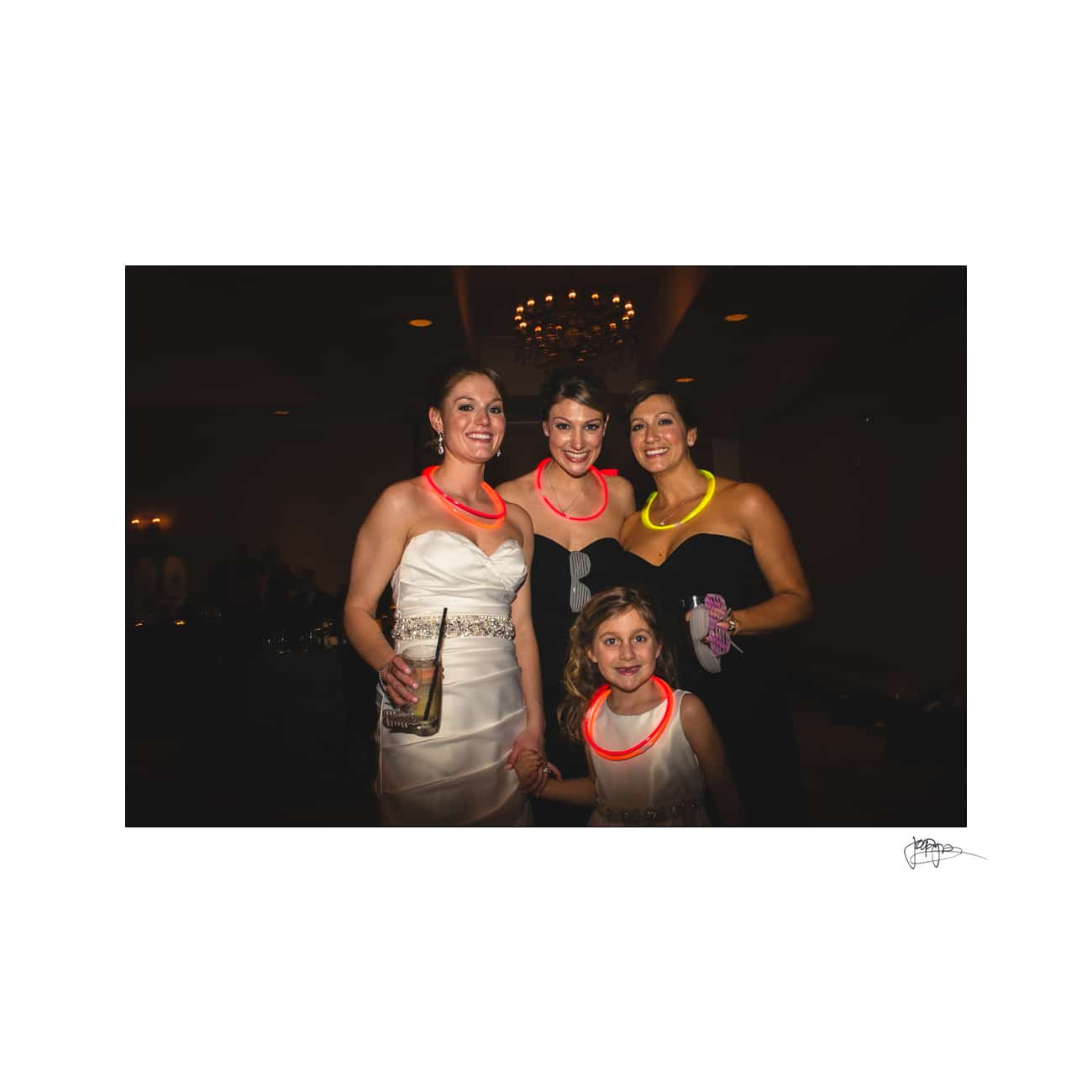 Four people at an indoor event, three adults wearing glow necklaces and holding drinks, and a young child in a white dress. The background is dimly lit with a chandelier visible above.
