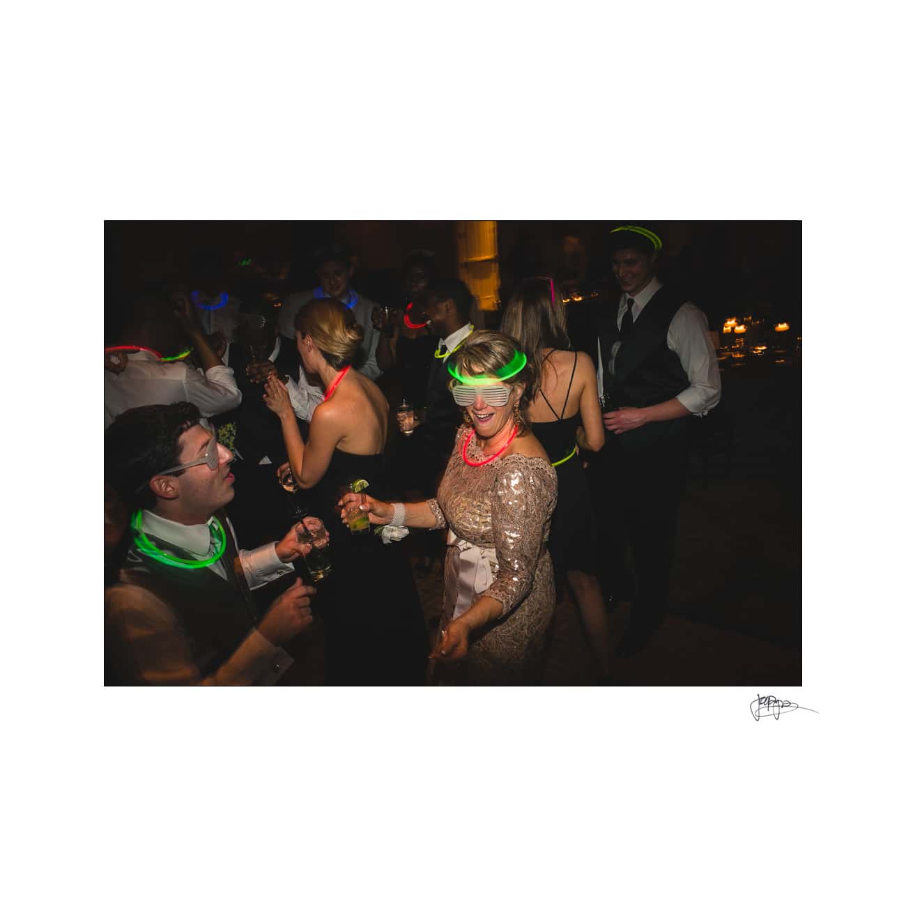 A lively party scene with people dancing and wearing glowing neon accessories. The focus is on a smiling woman in a gold dress, surrounded by others in formal attire enjoying the festive atmosphere.