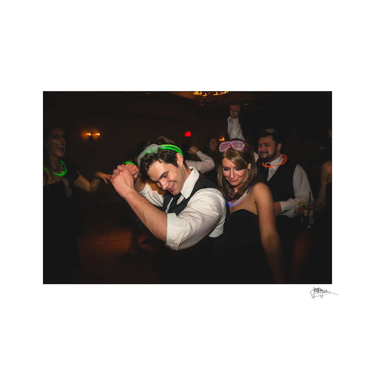 People dancing at a party, wearing glow stick necklaces and headbands. The man in the foreground wears a vest and shirt, while the woman has a strapless dress and pink shutter shades. They appear joyful, surrounded by other partygoers.