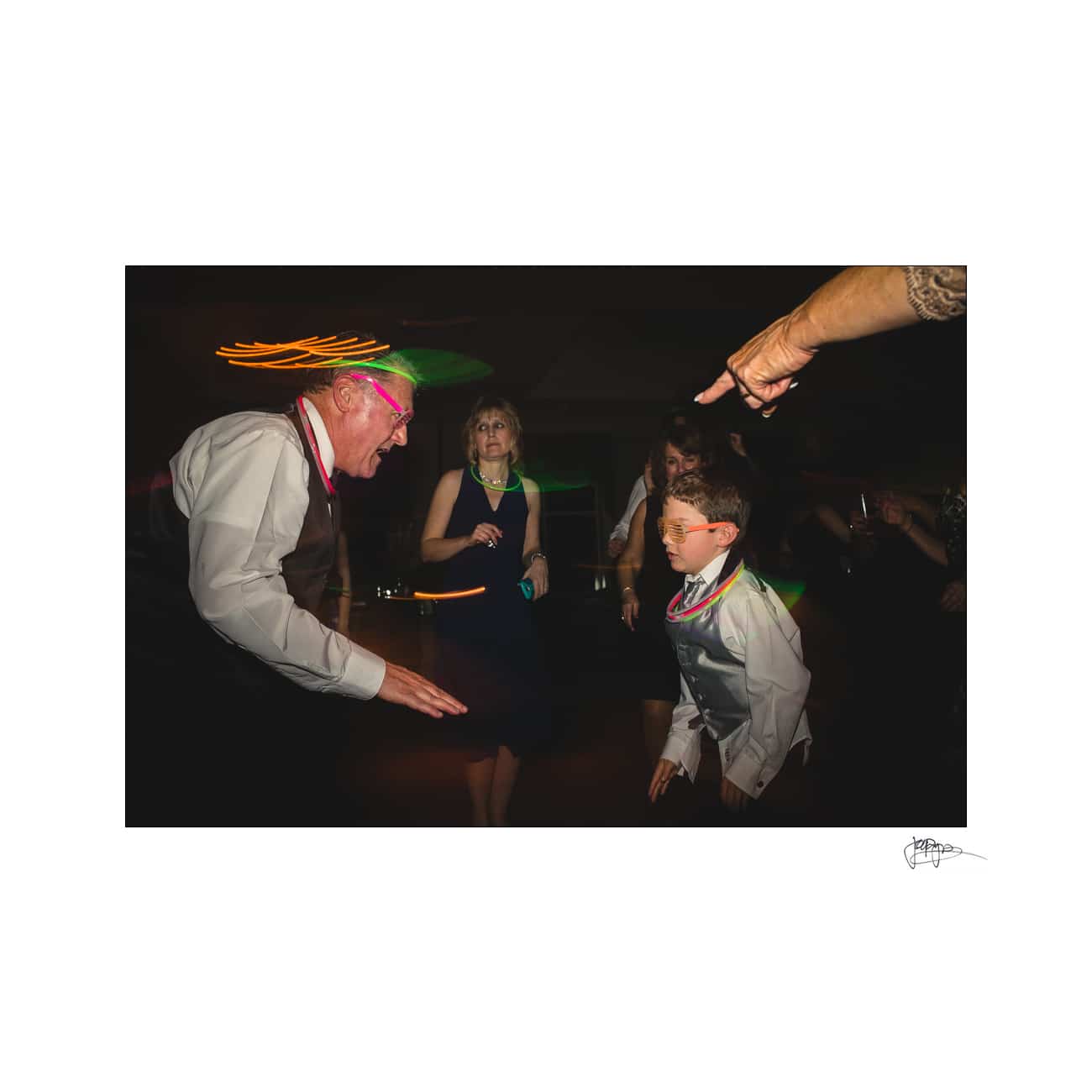 A lively dance floor scene with a man wearing a pink headband energetically dancing with a child in a suit and bow tie. A decorated hand points towards them, while a woman in black looks on, smiling in the background.