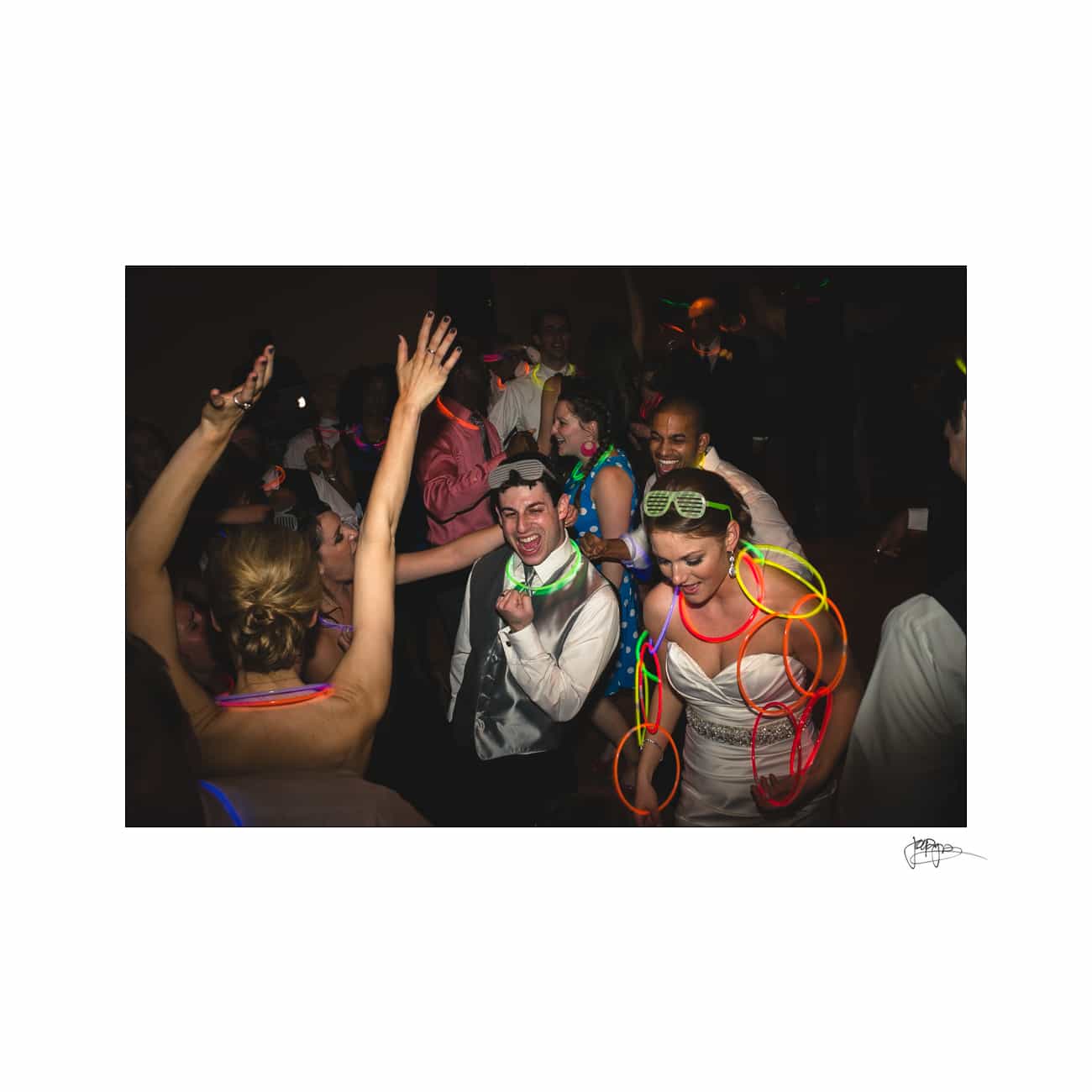 A group of people dancing at a party with colorful glow sticks. The atmosphere is lively, with smiling faces and diverse attire. The setting has dim lighting, accentuating the glow of the neon accessories on the partygoers.
