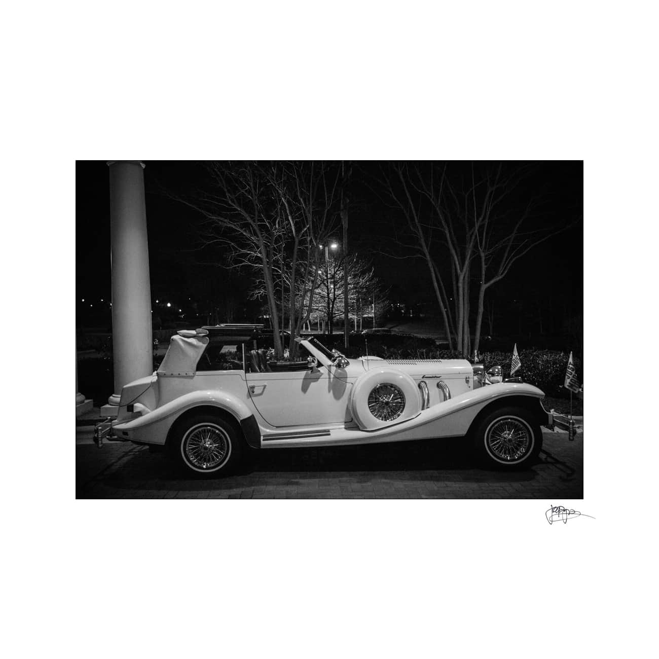 A vintage convertible car parked under a streetlight on a dark night. The black and white image highlights the classic design of the vehicle with its large fenders and spoked wheels. Bare trees and a column create a backdrop.