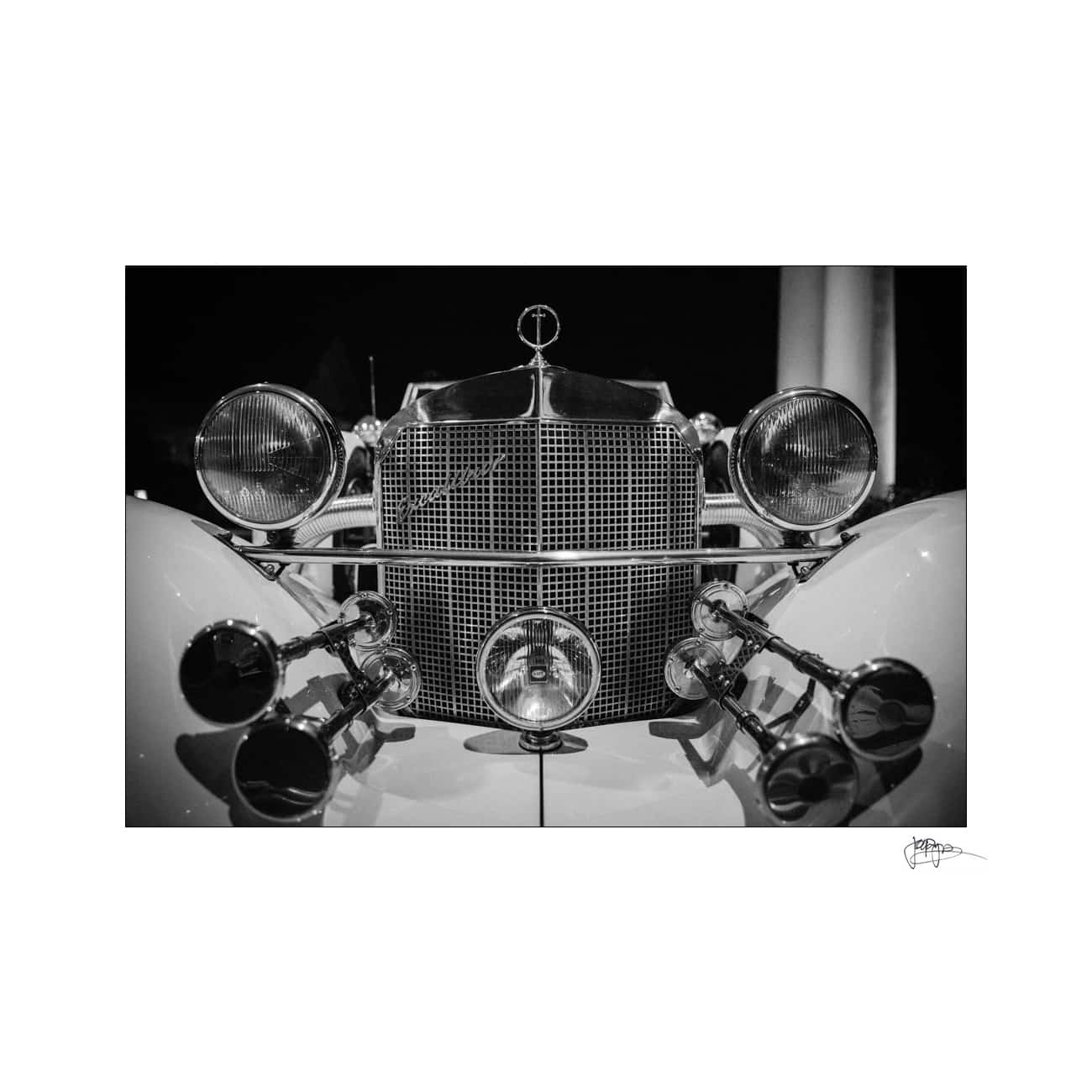 Black and white photo of a vintage cars front view, featuring a classic grille, round headlights, and an emblem on the hood. The intricate design showcases multiple chrome elements and a distinctive, elegant style.