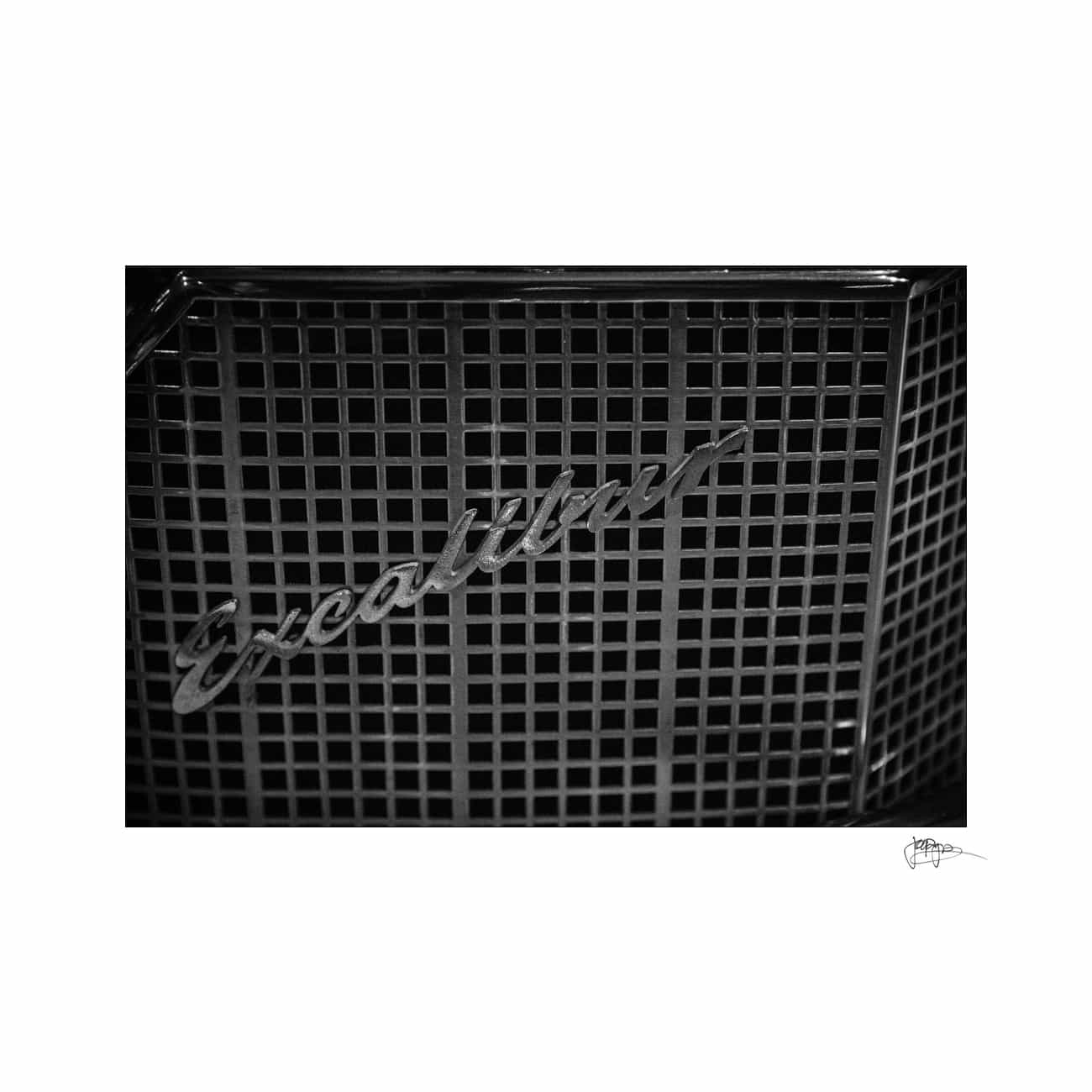 Black and white image of the front grille of a car with Excalibur written in cursive script. The grille has a mesh design.