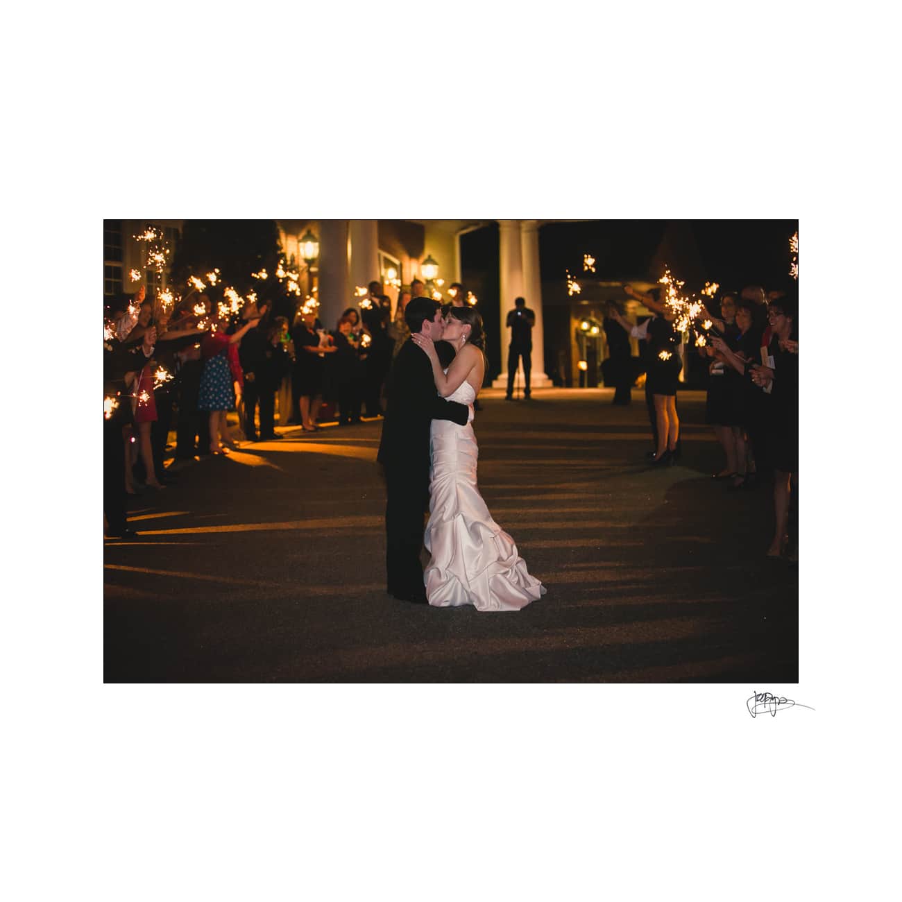 A bride and groom share a kiss in the center of a dimly lit area. They are surrounded by guests holding sparklers, creating a festive and romantic atmosphere.