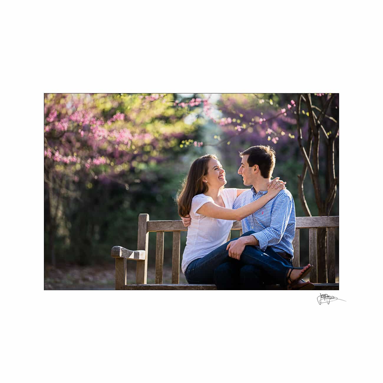 A couple sitting on a wooden bench outdoors, surrounded by blossoming trees, looking at each other and smiling. The sun is shining, creating a warm and cheerful atmosphere.