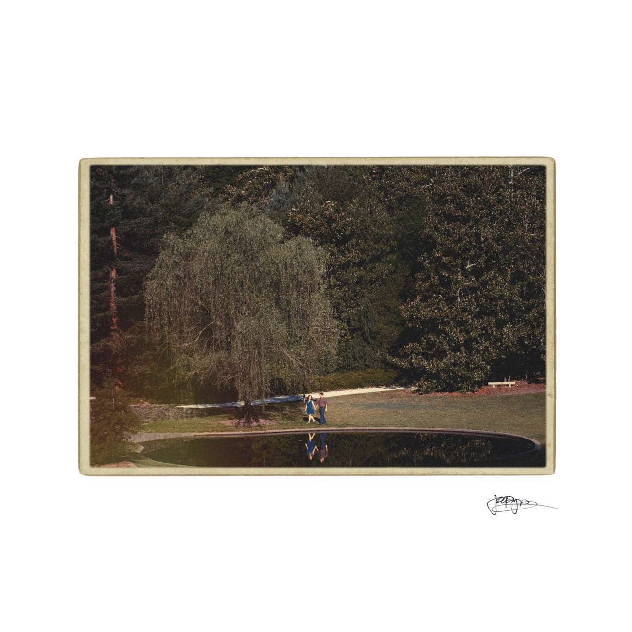 A vintage-style photo of two people walking beside a small pond in a park, surrounded by lush trees and greenery. A large willow tree stands out in the background.