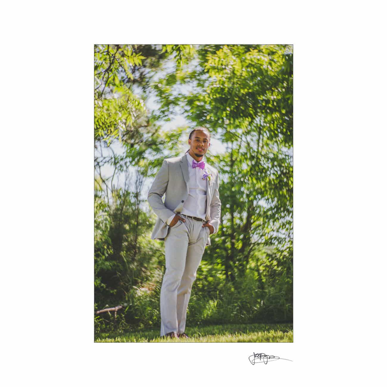 A man in a light gray suit with a magenta bow tie stands on grass, hands in pockets. Lush green trees and sunlight create a vibrant background. His expression is confident and relaxed.