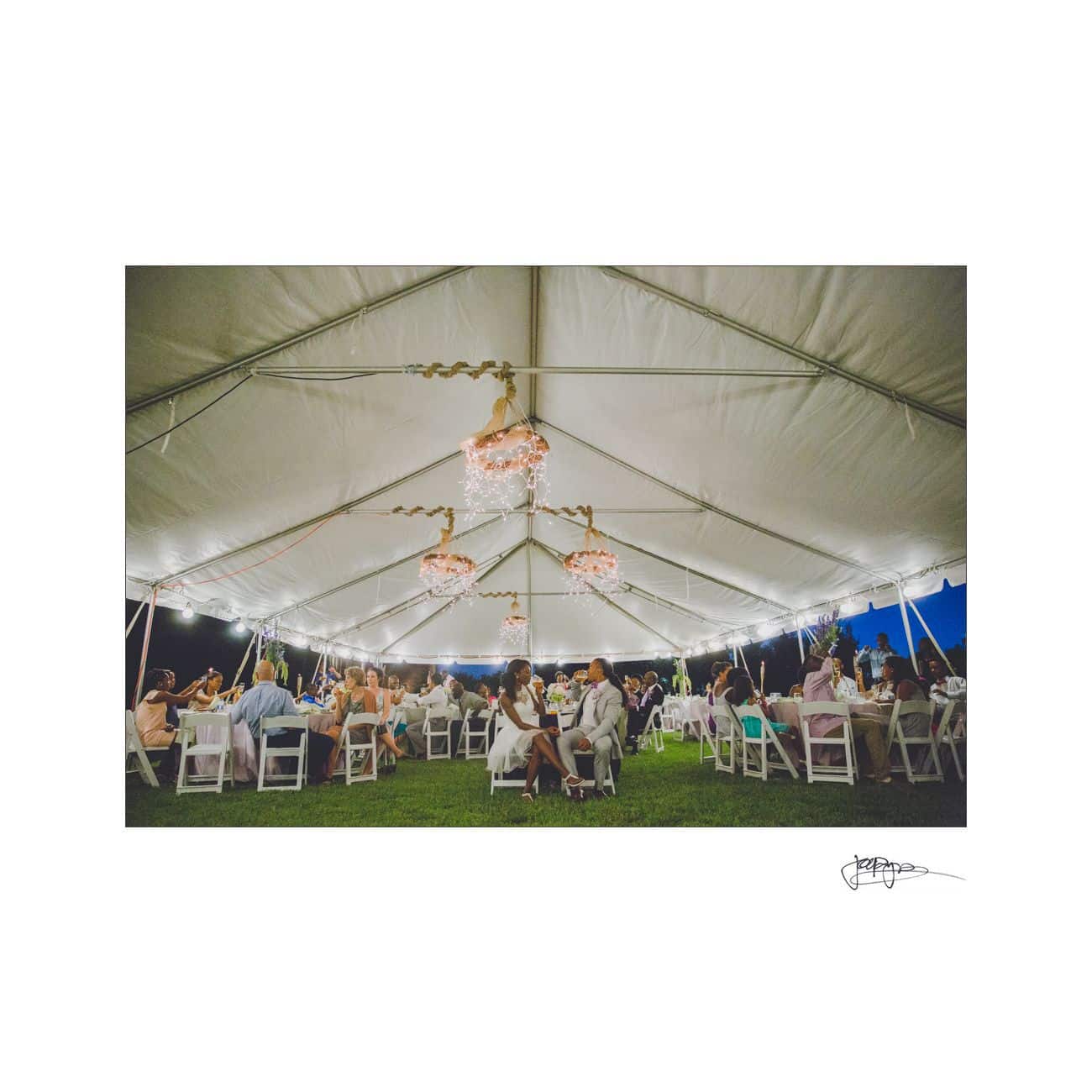 A wedding reception under a large white tent at night. Guests sit at round tables decorated with flowers. String lights hang above, creating a warm ambiance. Some people are chatting, while others enjoy their meals. Grass is visible outside the tent.