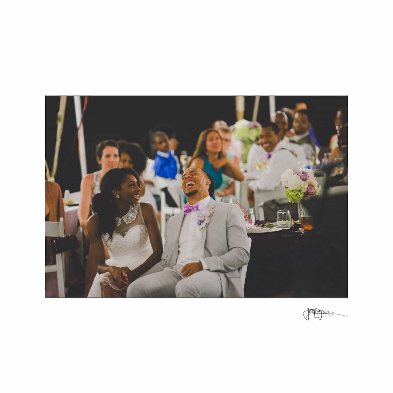 A bride and groom sit smiling and laughing among seated guests at an event. The groom wears a light gray suit and purple bow tie, and the bride is in a white dress. The atmosphere is joyful and festive.