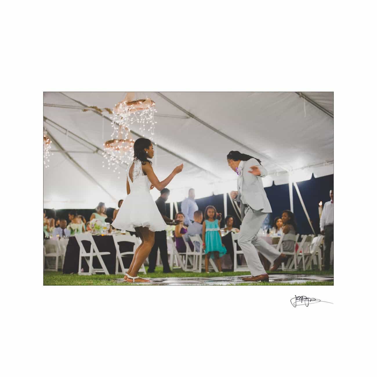 A couple dances energetically on a small stage under a white tent at an outdoor event. The woman wears a white dress, and the man is in a light suit. Guests sit at tables in the background, and lights hang from the tent ceiling.