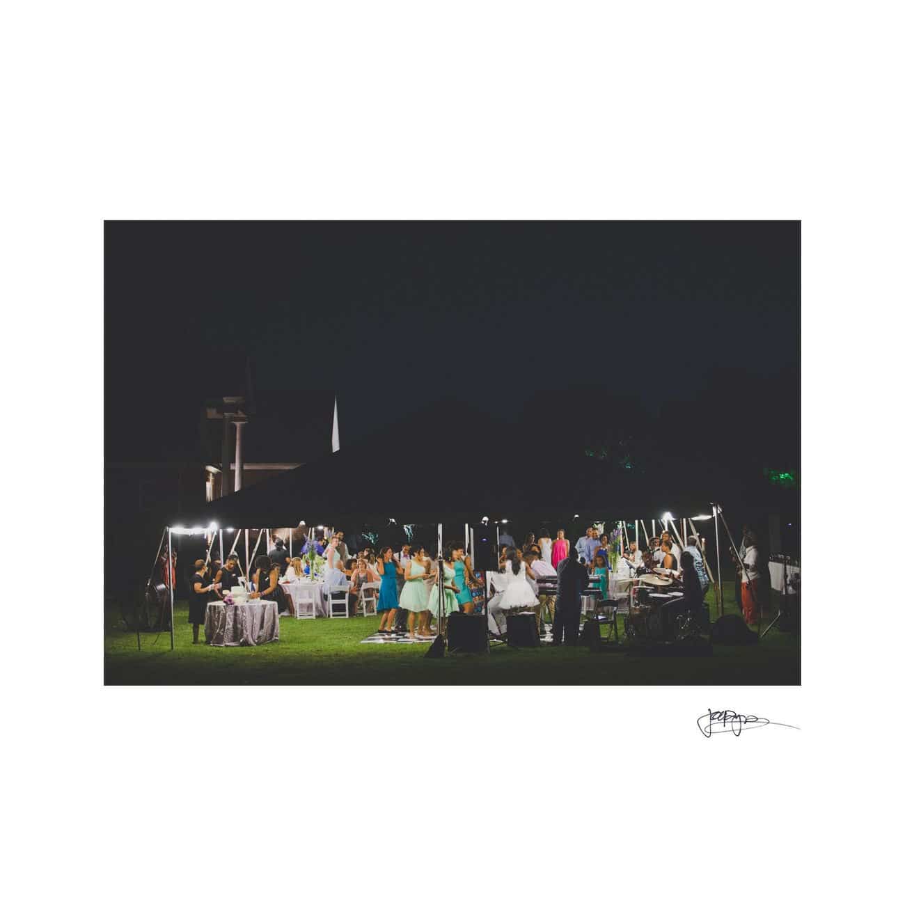 A vibrant outdoor gathering at night, with people mingling under string lights on a grassy area. Tables and chairs are set up, and the scene is filled with a lively, festive atmosphere.