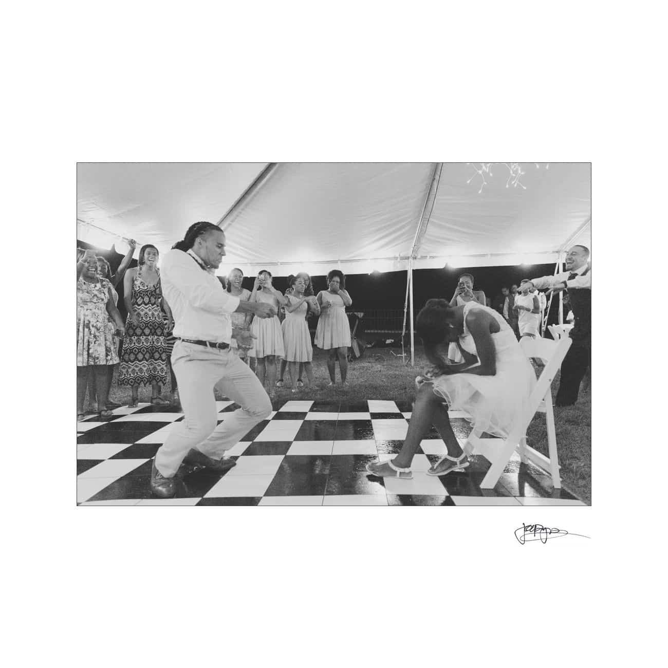 A person is playfully dancing in front of a seated person on a black-and-white checkered floor at a wedding reception. Guests are watching and smiling under a large tent.