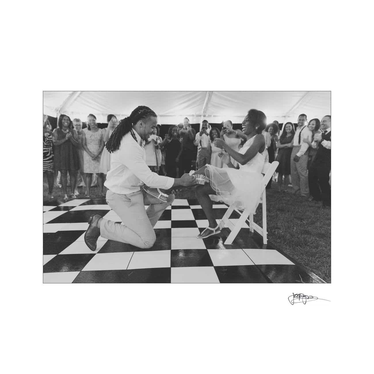 A person kneels on a checkered dance floor, playfully removing a garter from a seated partners leg. The couple is surrounded by smiling guests inside a tent, capturing a joyful moment during a wedding celebration.