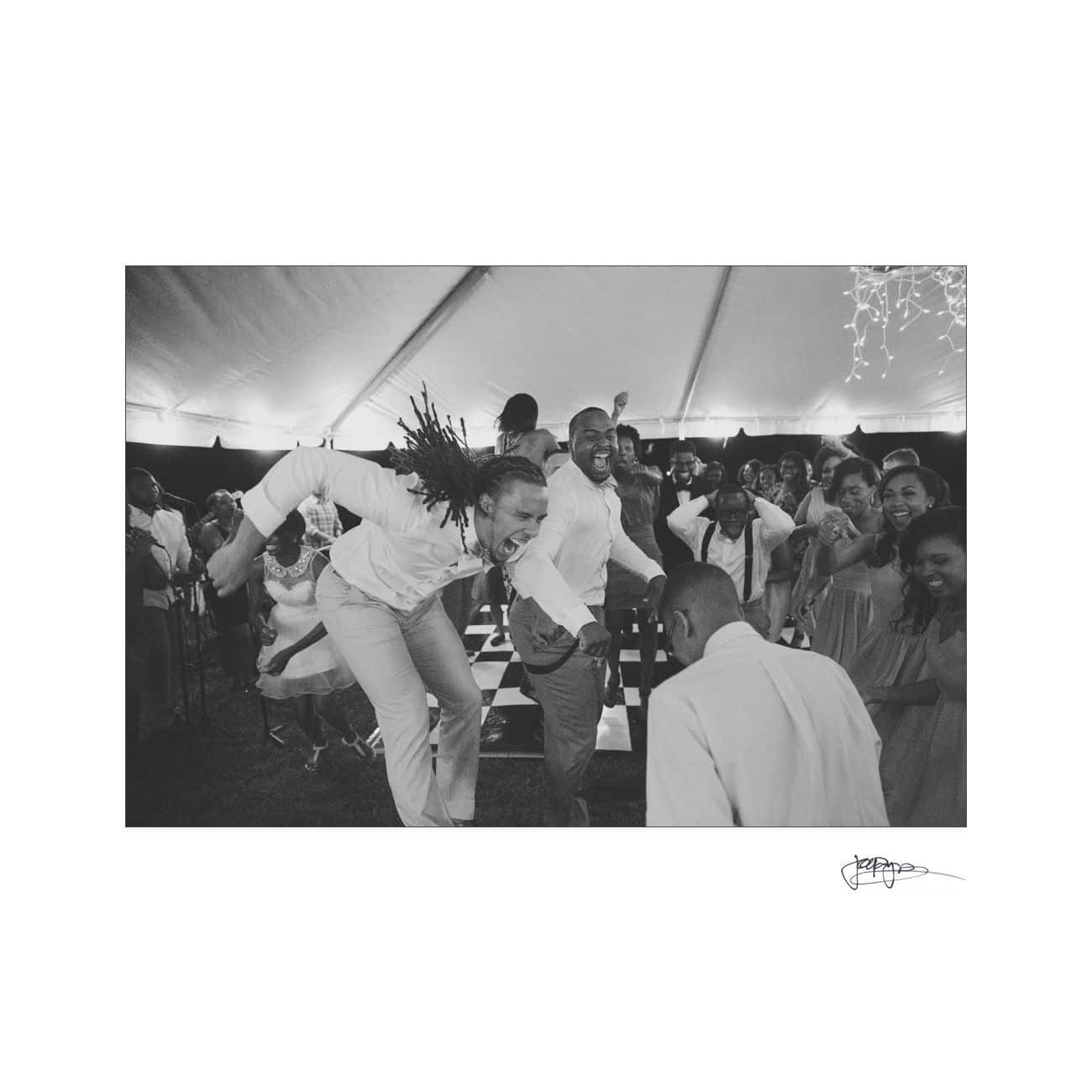A lively group of people dancing under a tent at a party. The atmosphere is joyful, with two individuals energetically leading the dance in the foreground, while others watch and join in the fun. The image is in black and white.