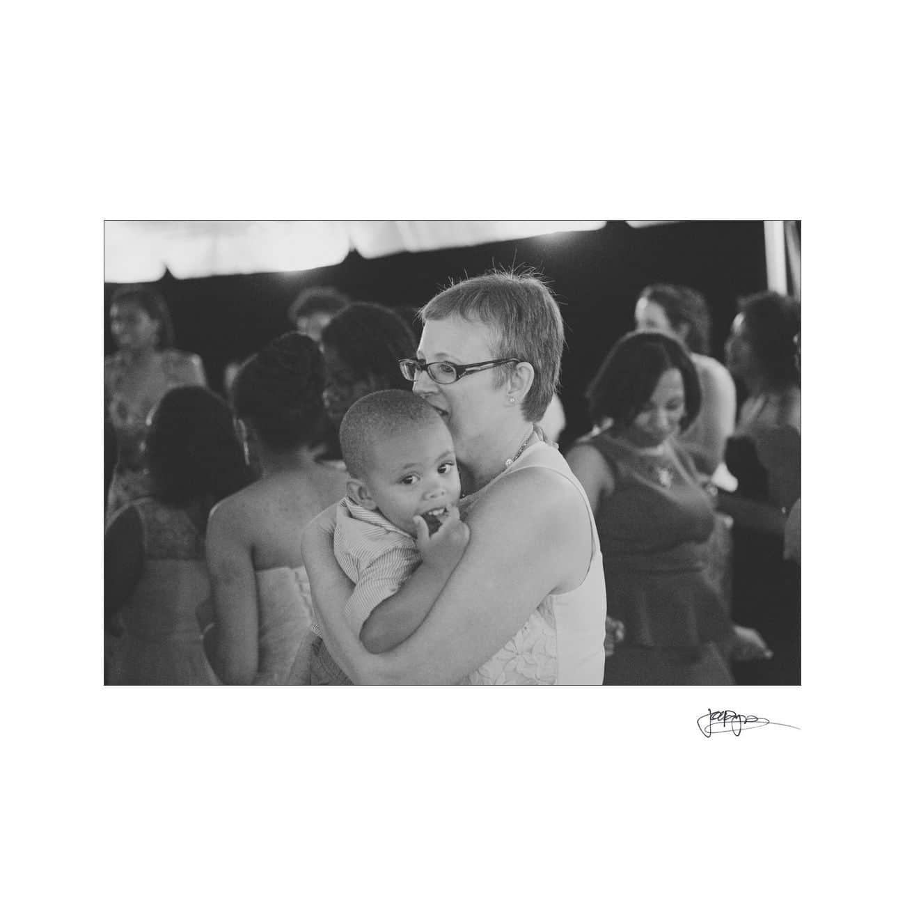 A black and white photo of a woman holding a young child at an event. The woman wears glasses and the child is looking curiously at the camera. People in the background are mingling under a tent.