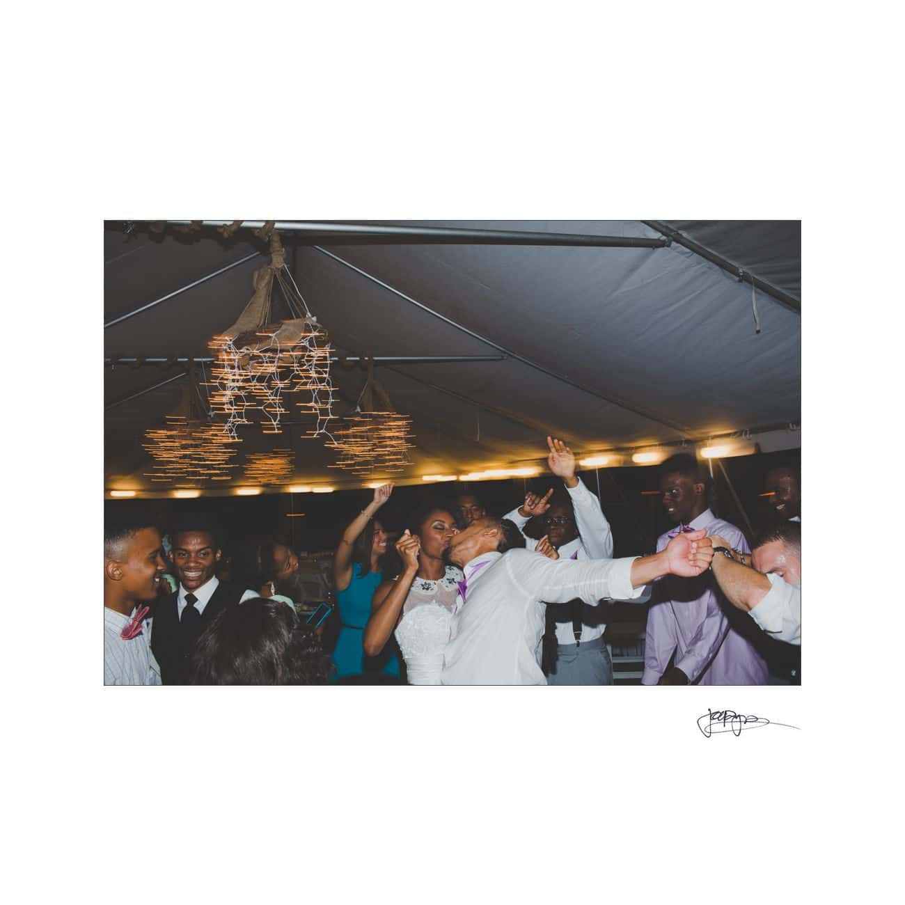 A joyful wedding reception with a newlywed couple kissing under a tent. Guests are celebrating, some with arms raised. Theres warm lighting and decorative hanging elements above. People wear formal attire in a festive atmosphere.