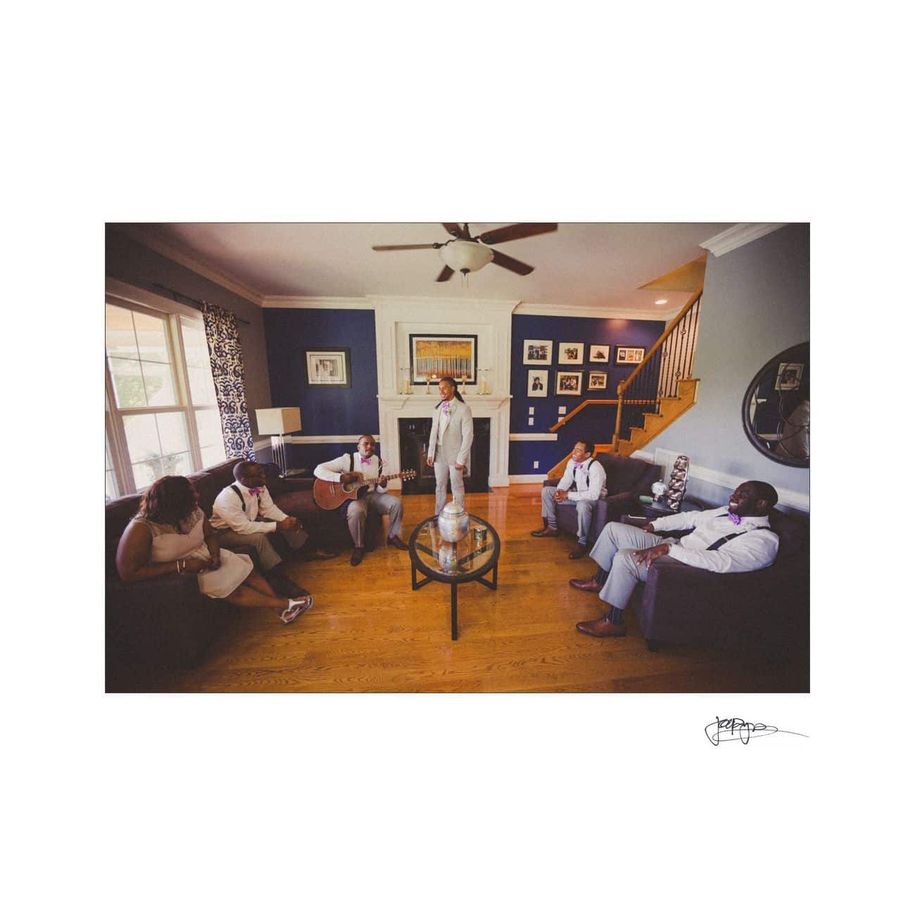 Five people in a living room are dressed in light suits, gathered around coffee tables. One person stands in the center, another plays guitar. The room has wooden floors, dark couches, and walls decorated with framed pictures.