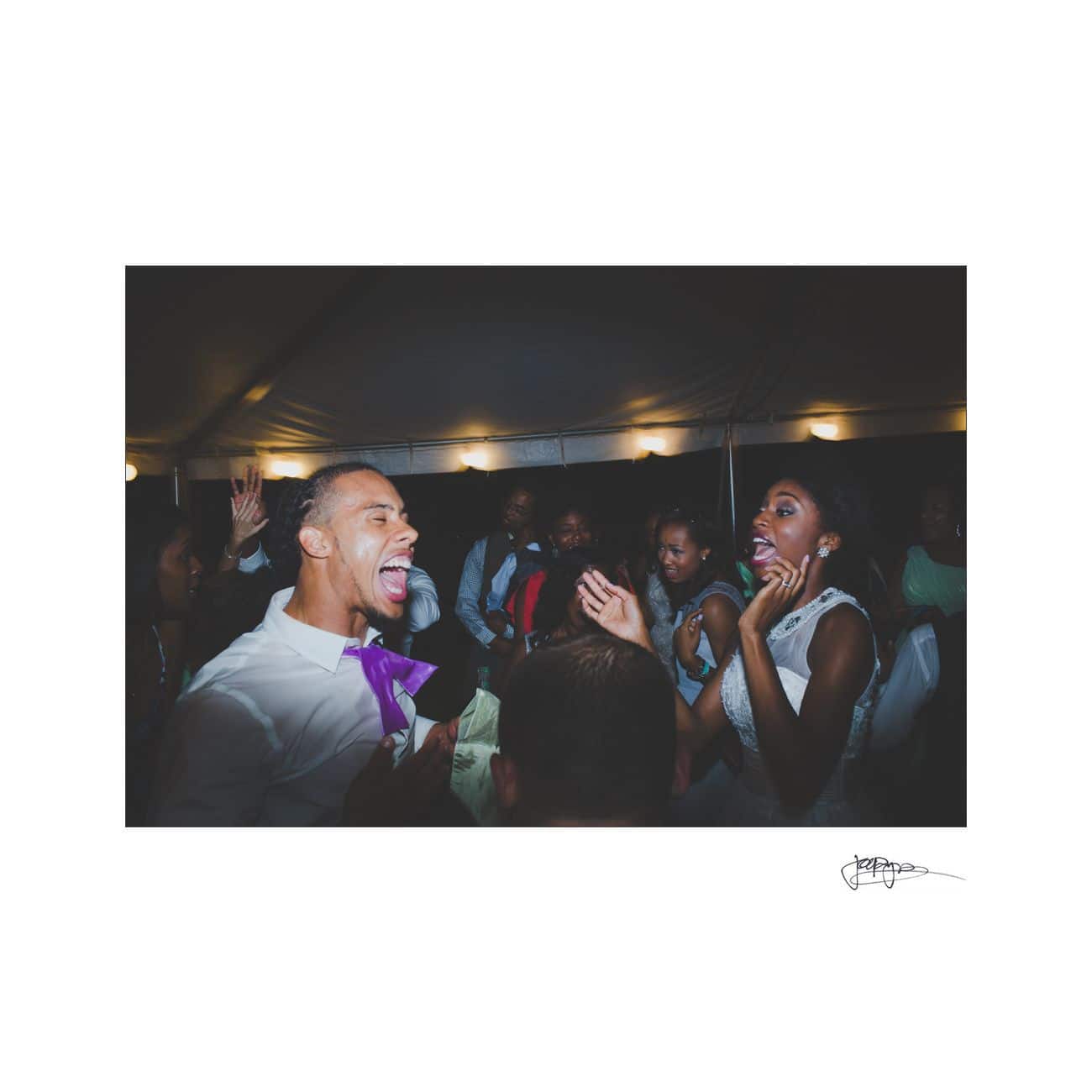 A group of people under a tent joyfully celebrating. A man in a white shirt and a woman in a white dress are at the center, laughing and cheering. The scene conveys a festive and lively atmosphere.