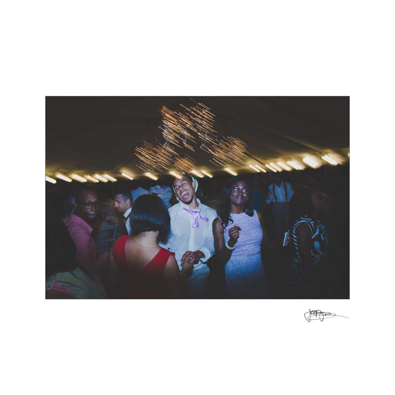 A lively party scene with people dancing, illuminated by dim lights. A man in a white shirt and pink bow tie is laughing joyously in the center. Blurry string lights hang above, adding a festive ambiance to the atmosphere.