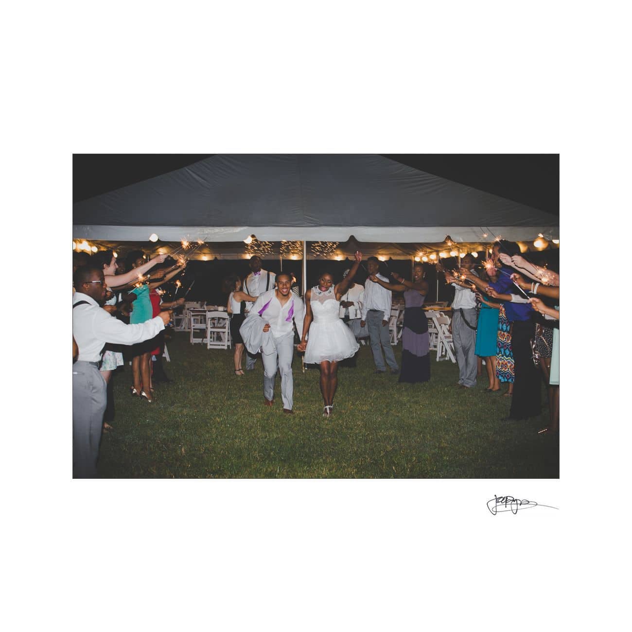 A joyful couple holding hands walks through a nighttime outdoor celebration. Guests are on either side, holding sparklers under a tent decorated with string lights. The scene conveys a festive, romantic atmosphere.