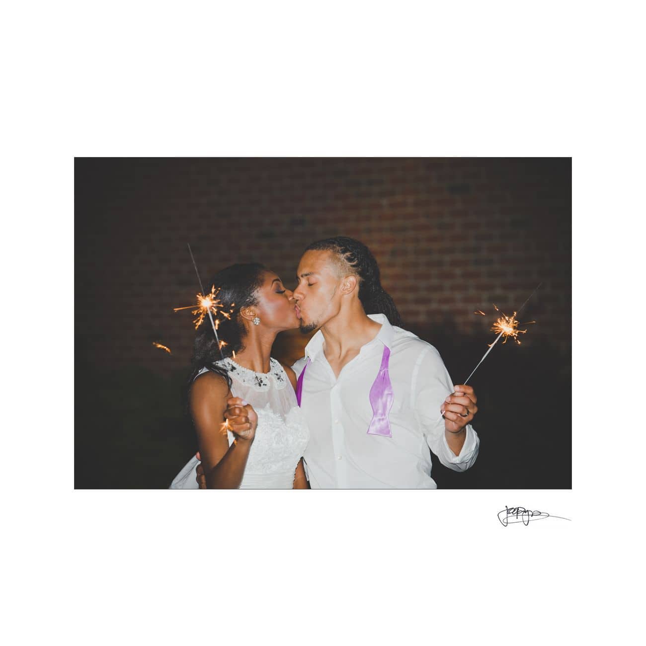 A couple kisses while holding sparklers at night. The woman wears a white dress and the man has a white shirt with a purple ribbon. They stand in front of a dark, blurred background.