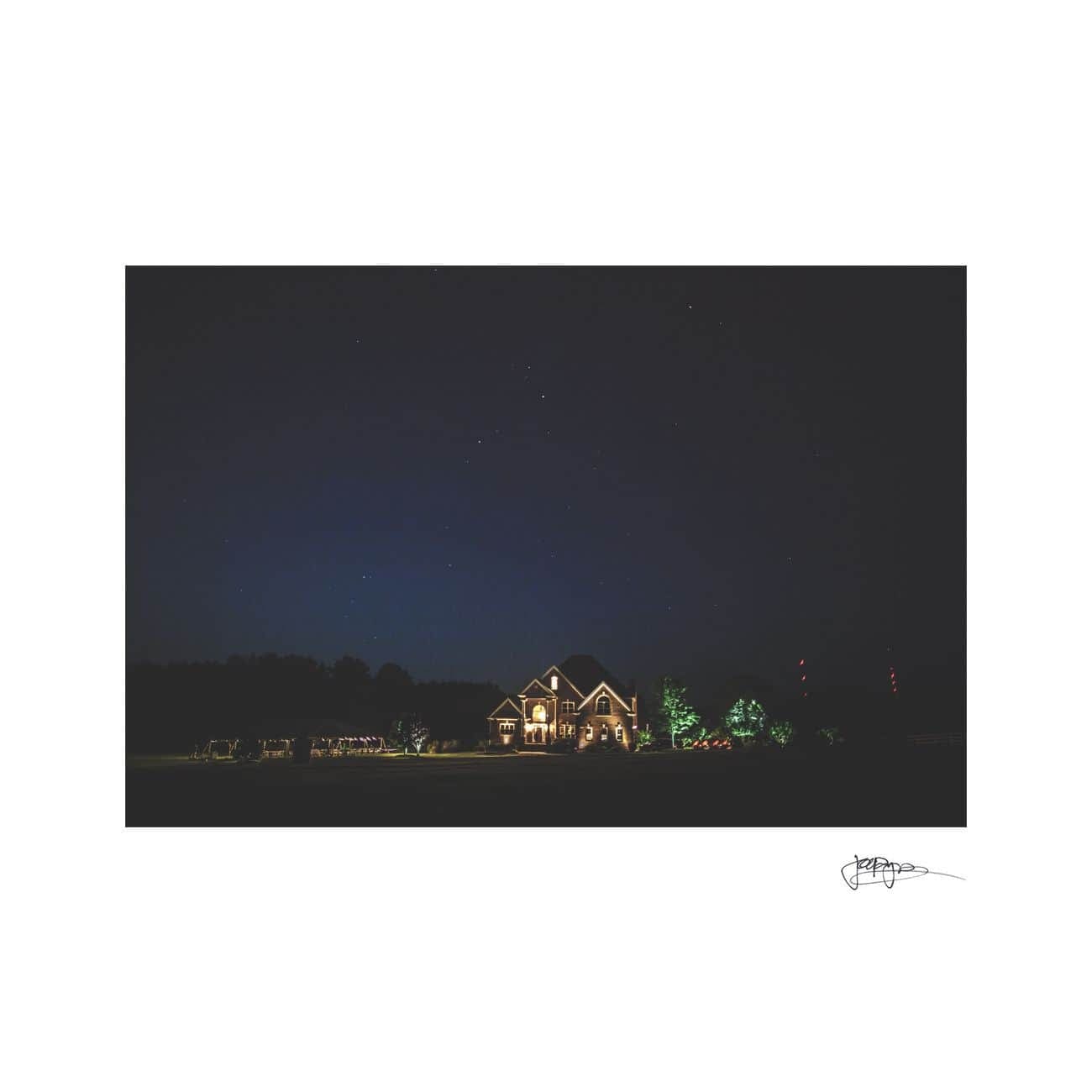 A large house with bright lights is surrounded by trees and an expansive lawn under a dark, starry sky. The house is centered in the image with a signature in the bottom right corner.