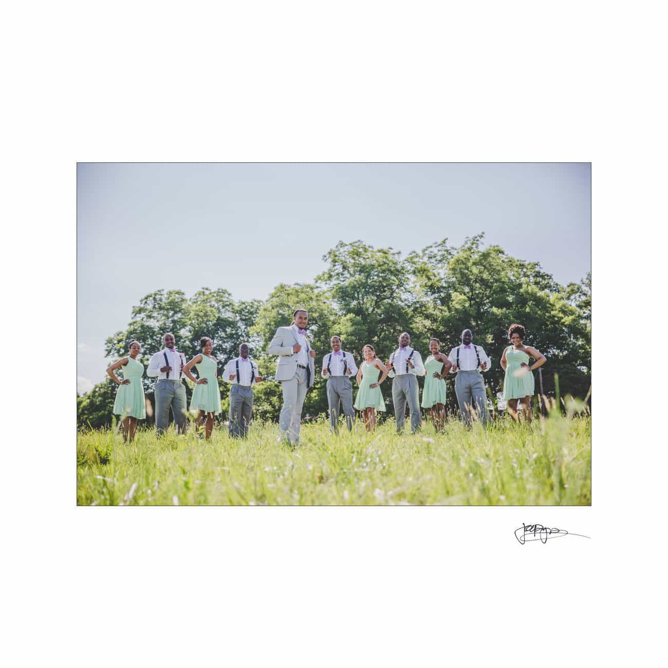 A group of people dressed in pastel-colored attire, standing in a field with green trees in the background. Some men are wearing suspenders. The sky is clear and sunny.