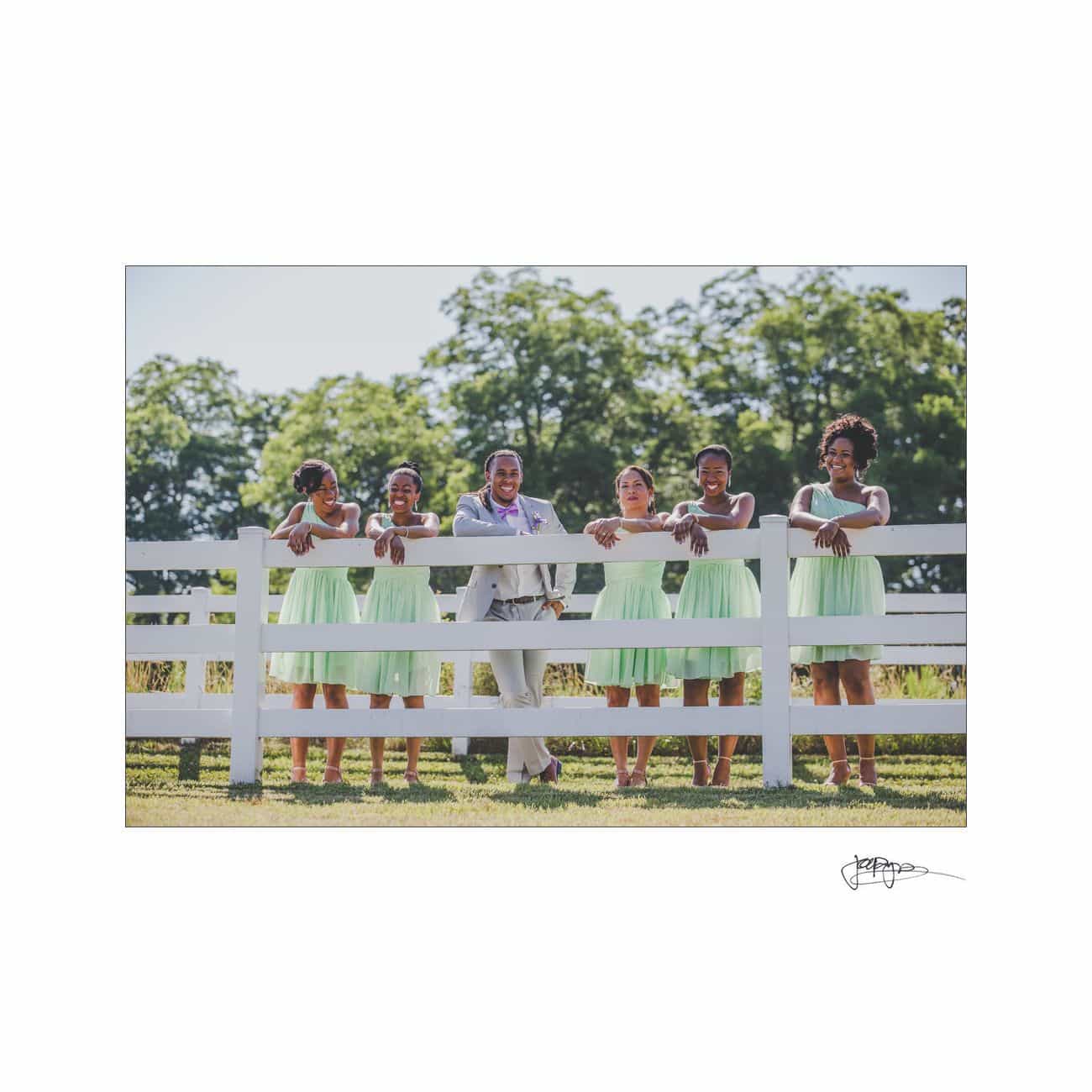 A group of people stand cheerfully behind a white fence. There is one person wearing a gray suit and five people in light green dresses. Trees and a clear sky are visible in the background.