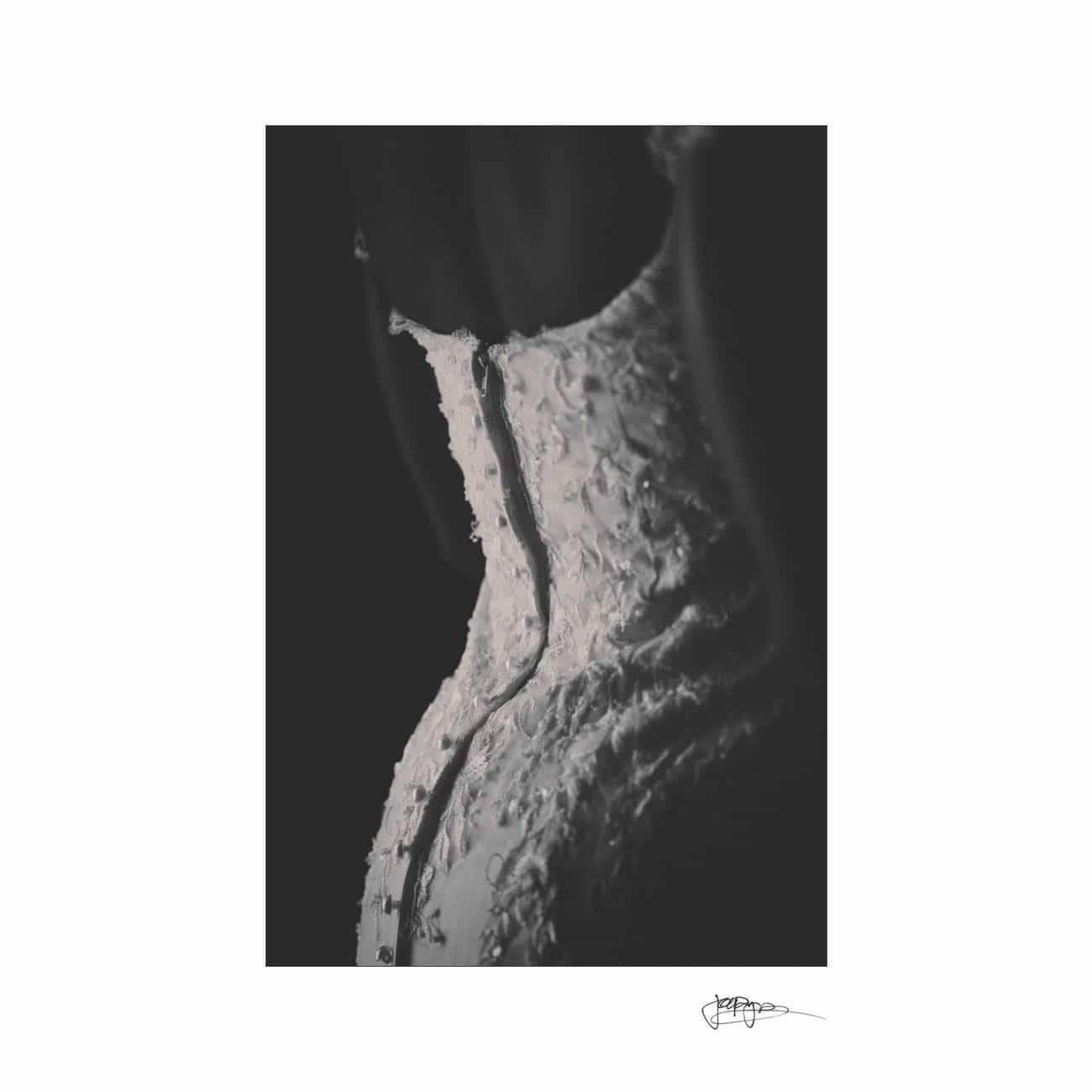 Black and white photo of a waistline in a detailed, textured garment with lace and floral patterns. The image focuses on the curves and intricate design of the fabric, with a signature at the bottom right corner.
