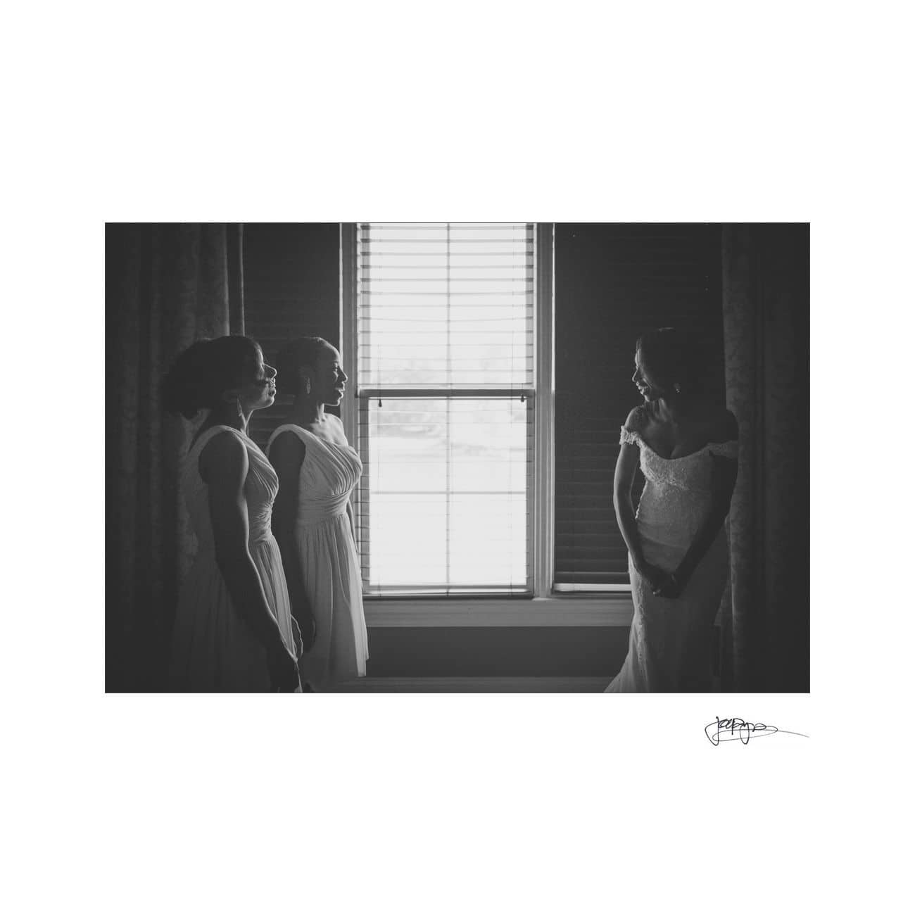 A bride in a wedding dress stands near a window, smiling at two bridesmaids. The room is softly lit, creating a silhouette effect. The scene is intimate and serene.