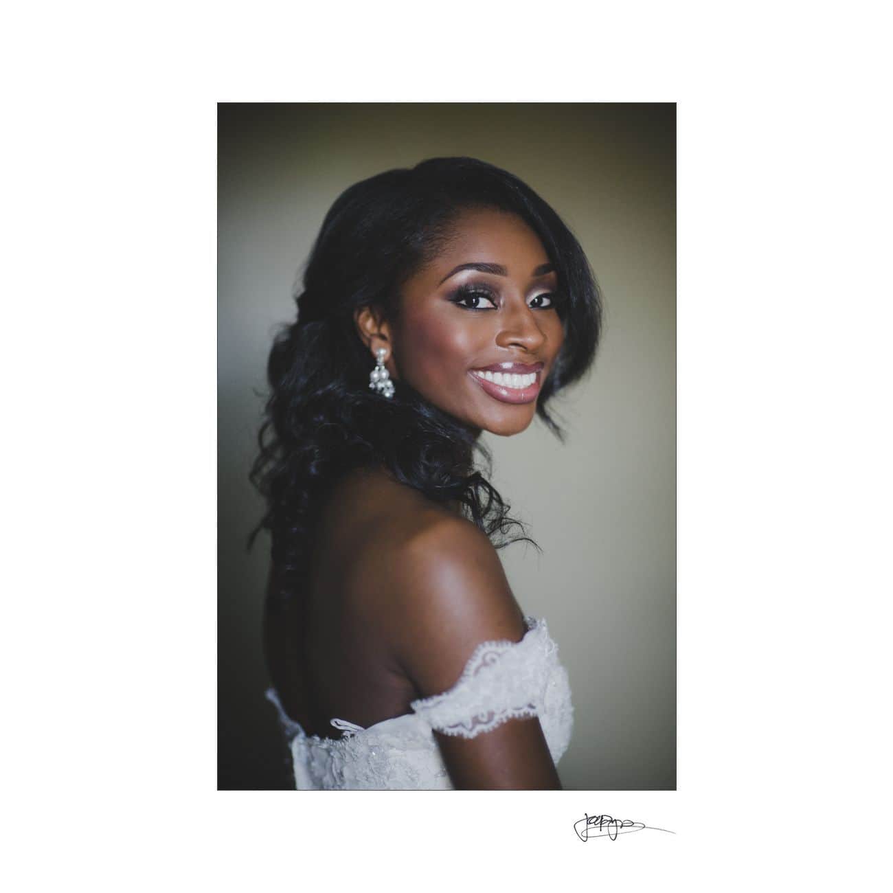 A woman in a wedding dress with off-the-shoulder lace details smiles warmly. She has long, wavy hair and wears drop earrings. The background is softly blurred, highlighting her joyful expression.