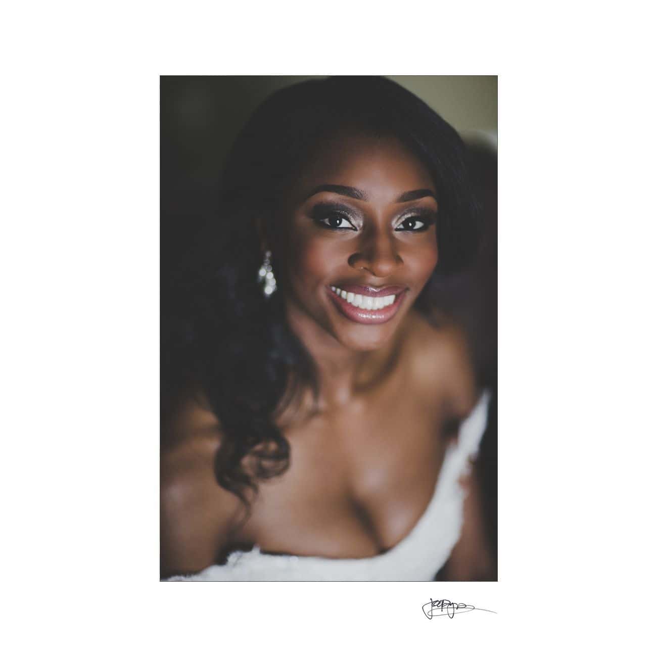 A woman in a bridal gown smiles warmly at the camera. She has long, dark hair and wears elegant earrings. The background is softly blurred, highlighting her joyful expression and the intricate details of her dress.